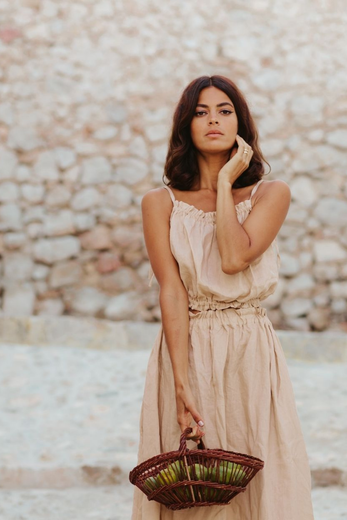 A woman with long brown hair stands outdoors in front of a stone wall, wearing a Mona Top by Myrah Penaloza. She holds a basket filled with green figs in one hand and touches her face with the other.