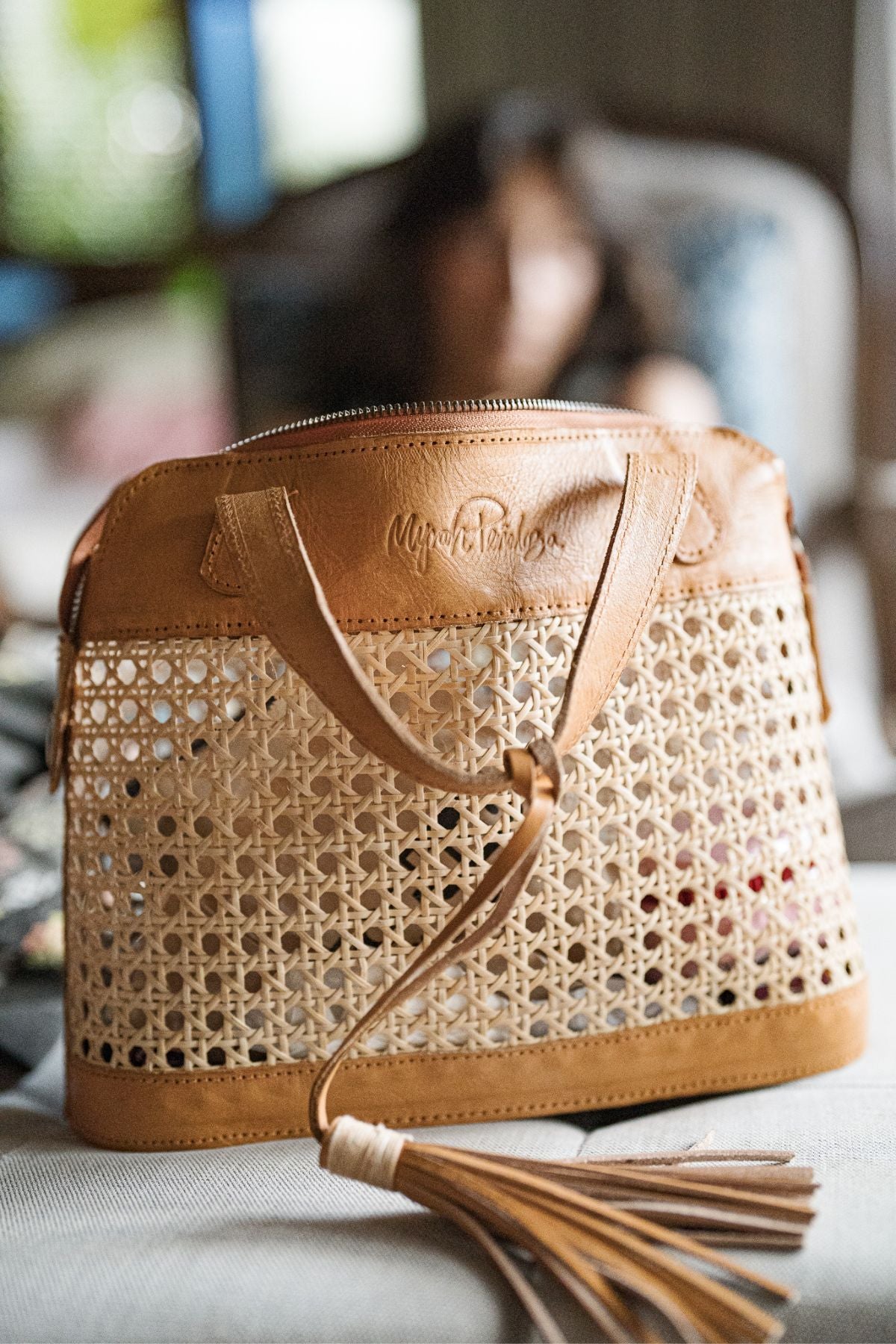 The Wonder Everyday Tote Mini by Myrah Penaloza is a medium-sized handcrafted Bali tote made of tan leather and natural plant fibers, featuring a leather handle and detailed tassels. The bag boasts an embossed logo on the leather portion and sits on a softly lit surface with a blurred background.