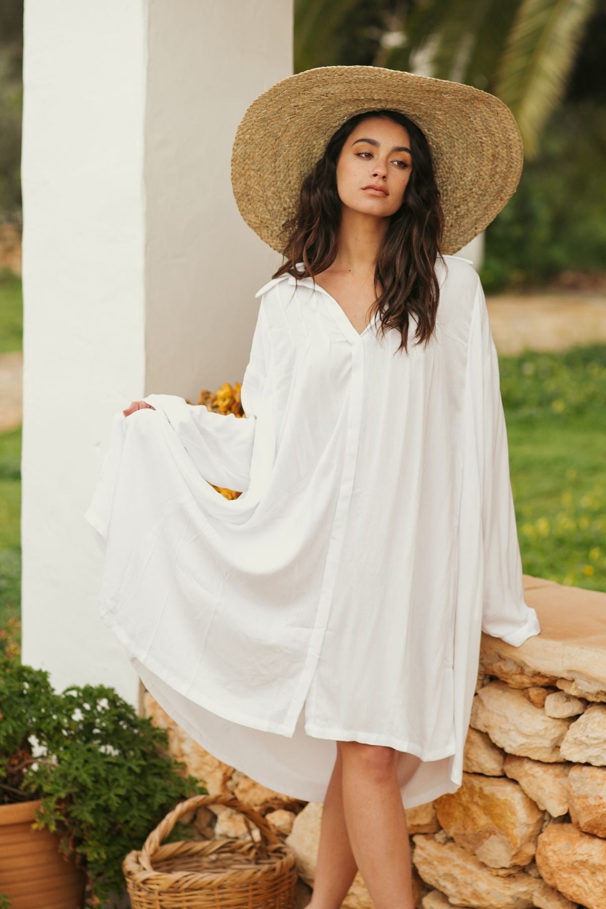 A woman dressed in an off-white Kundalini Gown Short, made from 100% Bamboo Rayon by Myrah Penaloza, and wearing a wide-brimmed straw hat stands by a stone wall. She gently holds the gown's fabric with one hand outdoors, surrounded by greenery and a pillar in the backdrop. Nearby, a wicker basket overflowing with plants complements the natural appeal of this handmade garment.