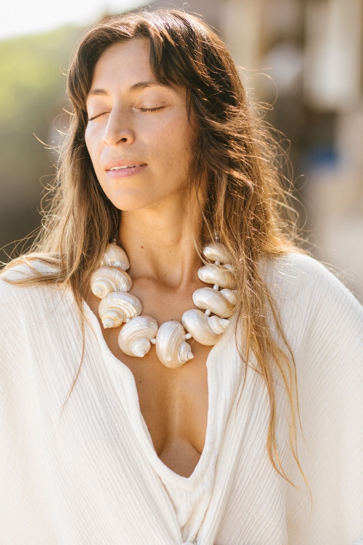 A woman stands outdoors with her eyes closed, wearing a stunning large Aphrodite's Treasure Necklace from Myrah Penaloza and a flowing white garment. Her long, wavy hair cascades over her shoulders, beautifully merging into the softly blurred background.