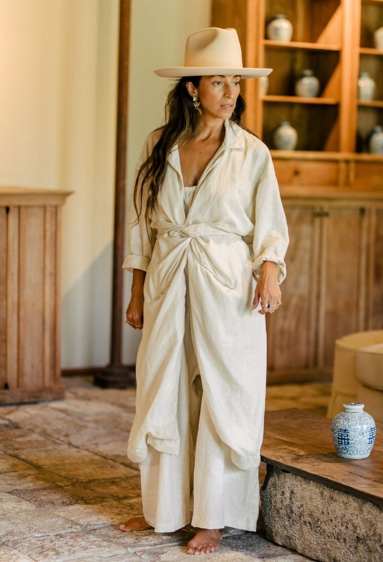 A woman stands in a room with wooden furniture, wearing an Infinity Jacket from Myrah Penaloza and a wide-brimmed hat. Her loose, tied beige outfit is made entirely of linen. She is barefoot on a textured floor, alongside a decorative blue and white ceramic vase.