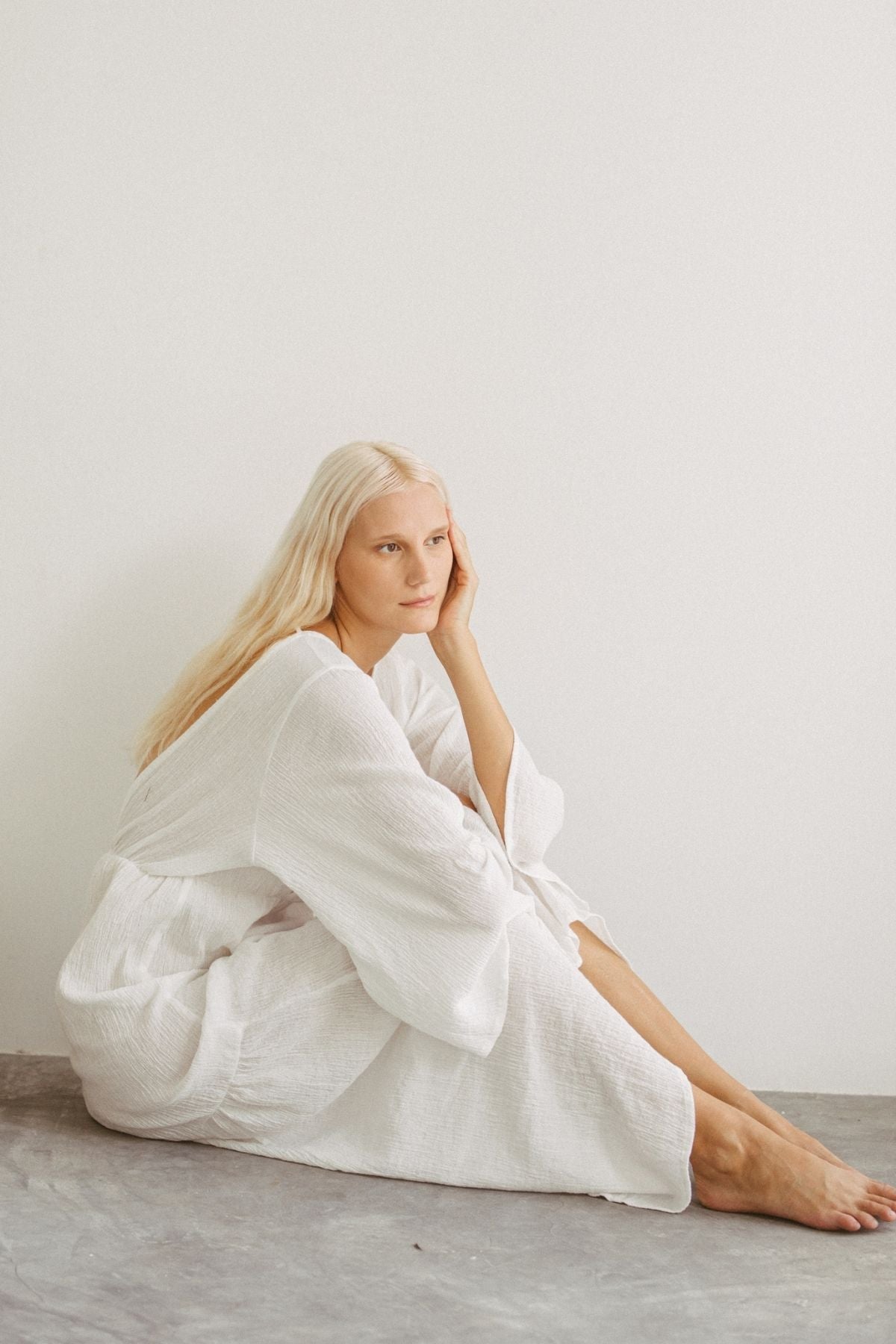 A woman with long blonde hair sits on the floor, leaning against a white wall. She is wearing the elegant Sayan Gown (Linen/Cotton) by Myrah Penaloza and rests her chin on her hand, gazing thoughtfully into the distance. The scene is minimalistic, exuding a calm and serene atmosphere.