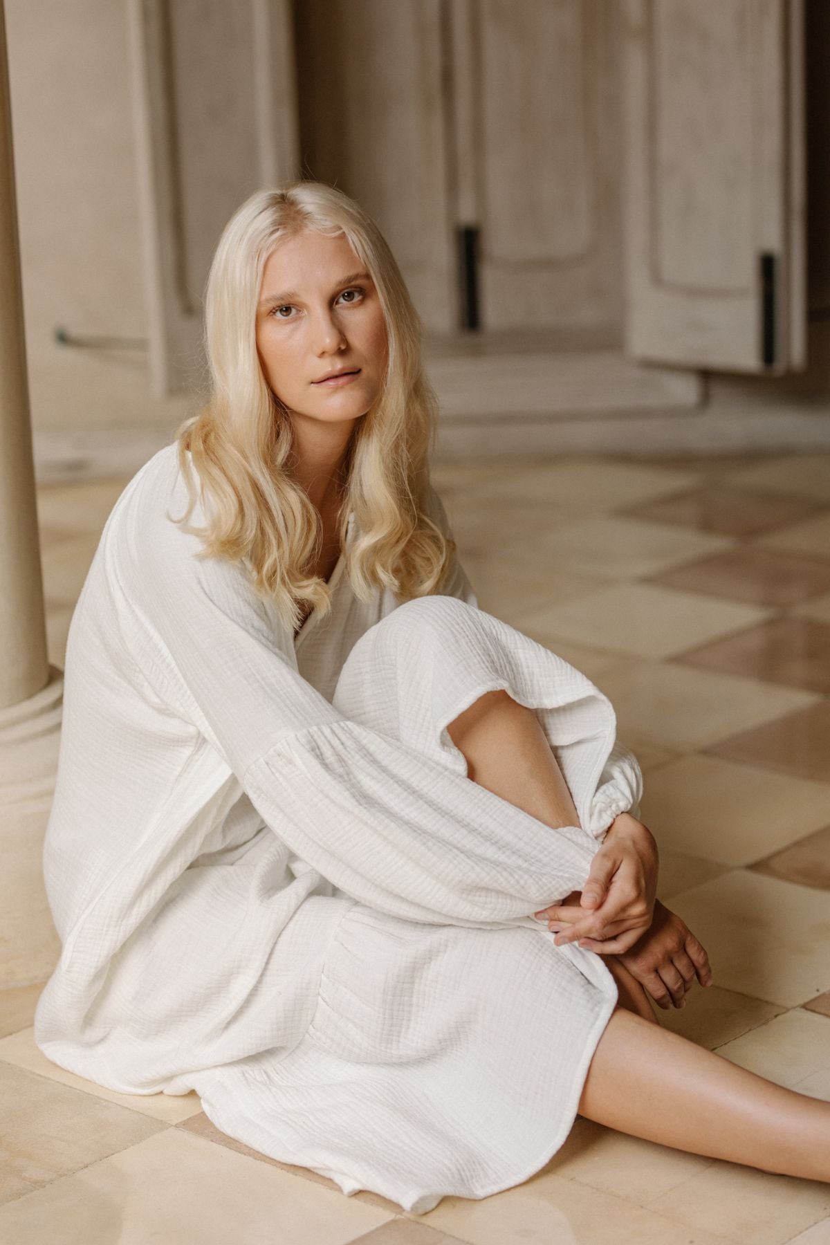 A woman with long blonde hair sits on the floor in a relaxed pose, wearing the Venus Gown - Cotton Double Deo by Myrah Penaloza. The background features light-colored, polished flooring and distressed, white wooden doors. Exuding comfort, she looks directly at the camera with a calm expression.