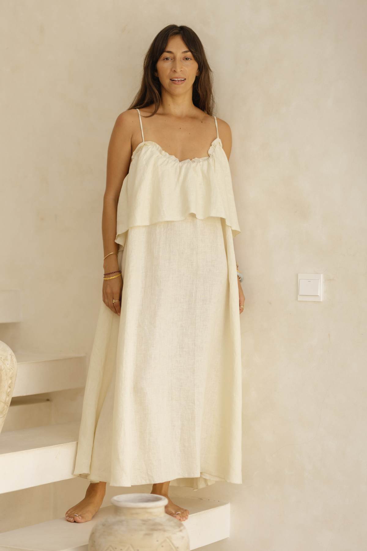 A woman with long, dark hair stands on a staircase against a neutral background. She is wearing the Linen Muse Gown by Myrah Penaloza, a long, cream-colored, sleeveless dress with adjustable straps and a ruffled top. Large decorative vases are positioned near the stairs. She looks towards the camera and smiles softly.