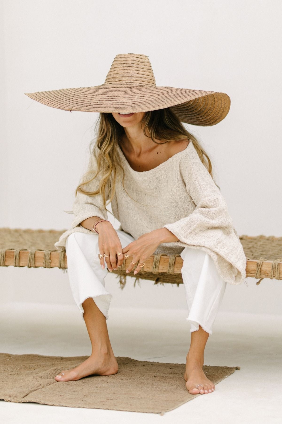 A woman with long hair sits on a woven bench, wearing a wide-brimmed straw hat that obscures her face, a loose beige top, and comfortable Miley High Waisted Jeans from Myrah Penaloza. She is barefoot, with one leg crossed over the other and hands clasped in front of her. The background is plain and minimalistic.