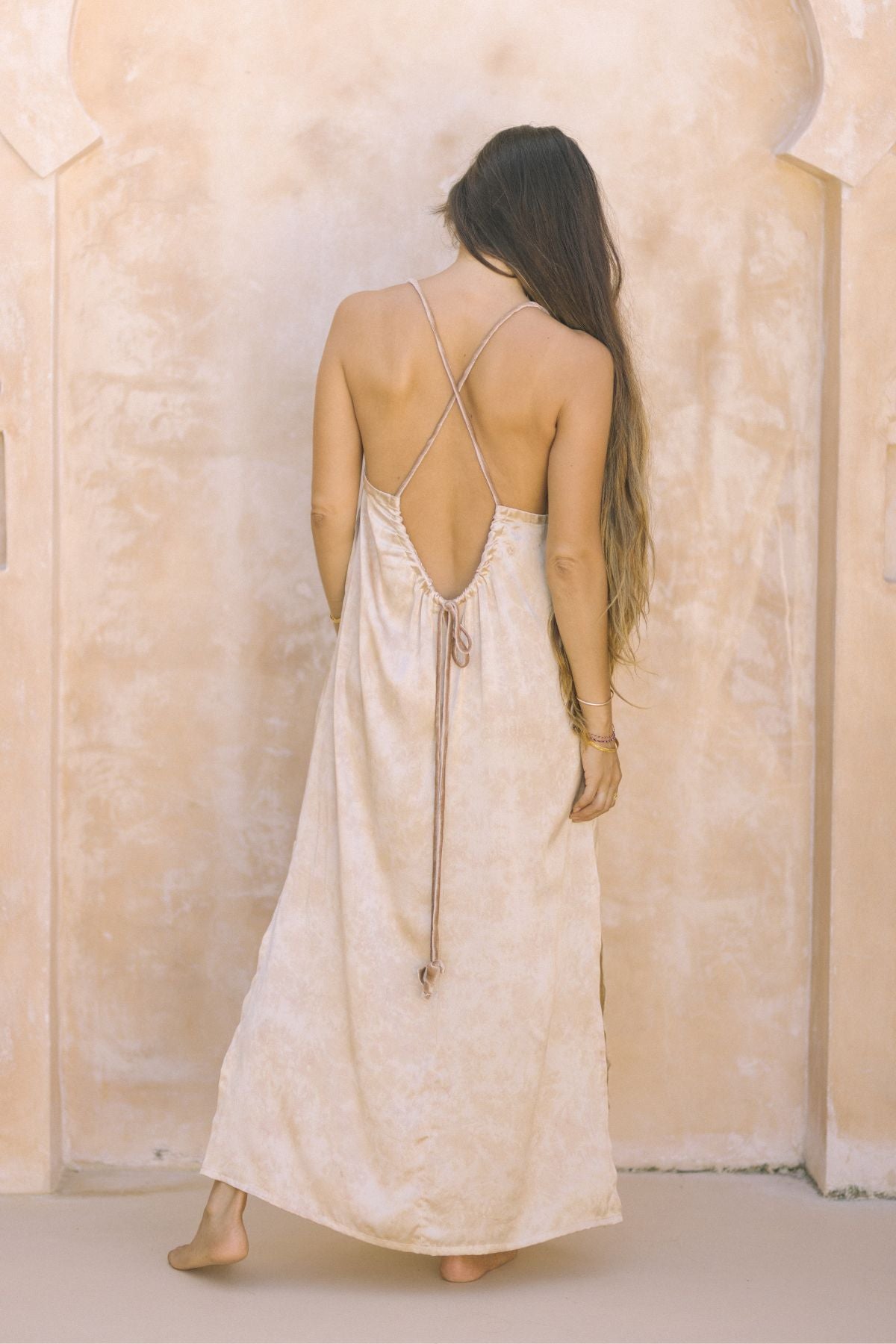 A woman with long hair, standing barefoot and facing away, wears the Gather Slip from Myrah Penaloza— a luxurious, backless dress crafted from 100% silk and botanically dyed rose. The scene is set against a textured, beige wall.