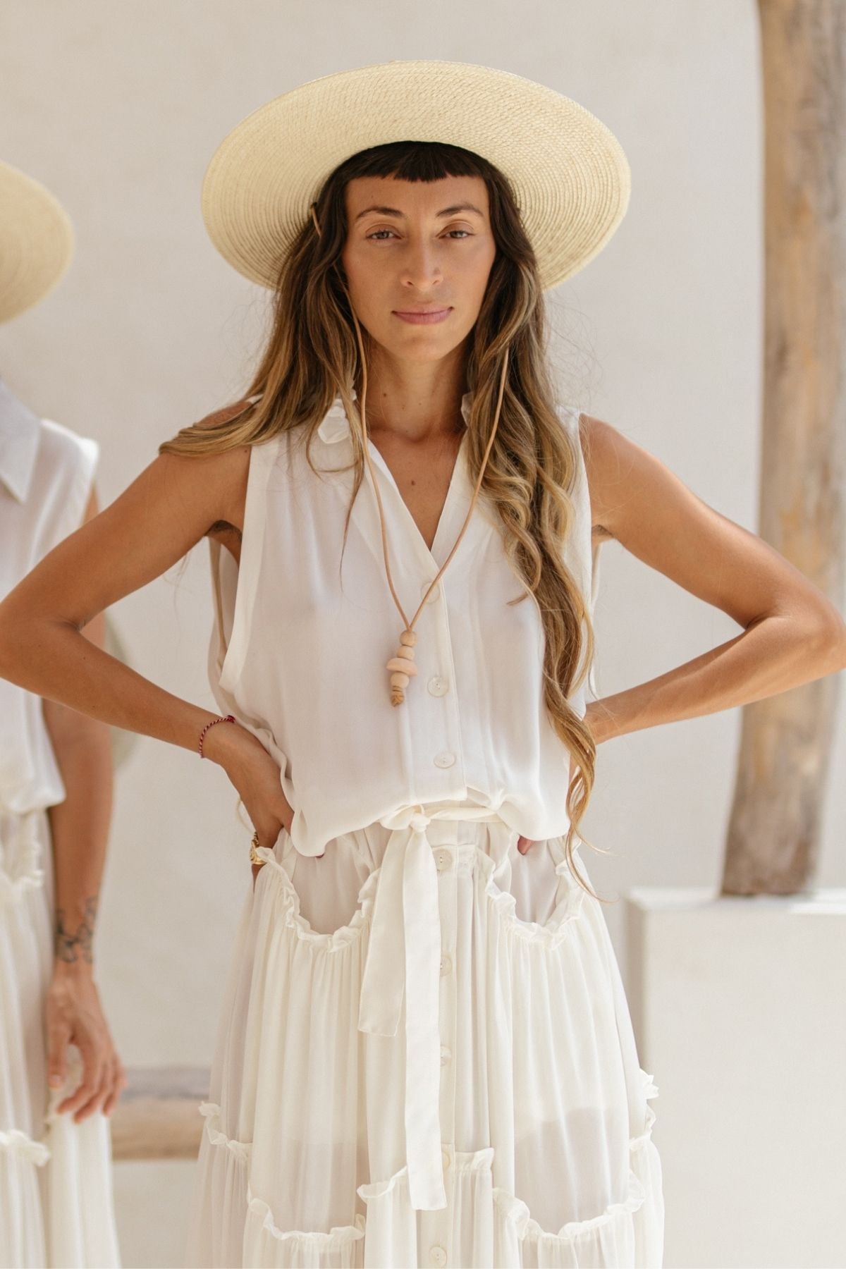 A woman in a La Dama by Myrah Penaloza, a sleeveless ruffle neck dress crafted from 100% silk, paired with a wide-brimmed hat reminiscent of a modern La Dama, stands confidently with her hands on her hips. Her long hair cascades over her shoulders, while a pendant necklace completes the look. The softly blurred background adds an ethereal touch.