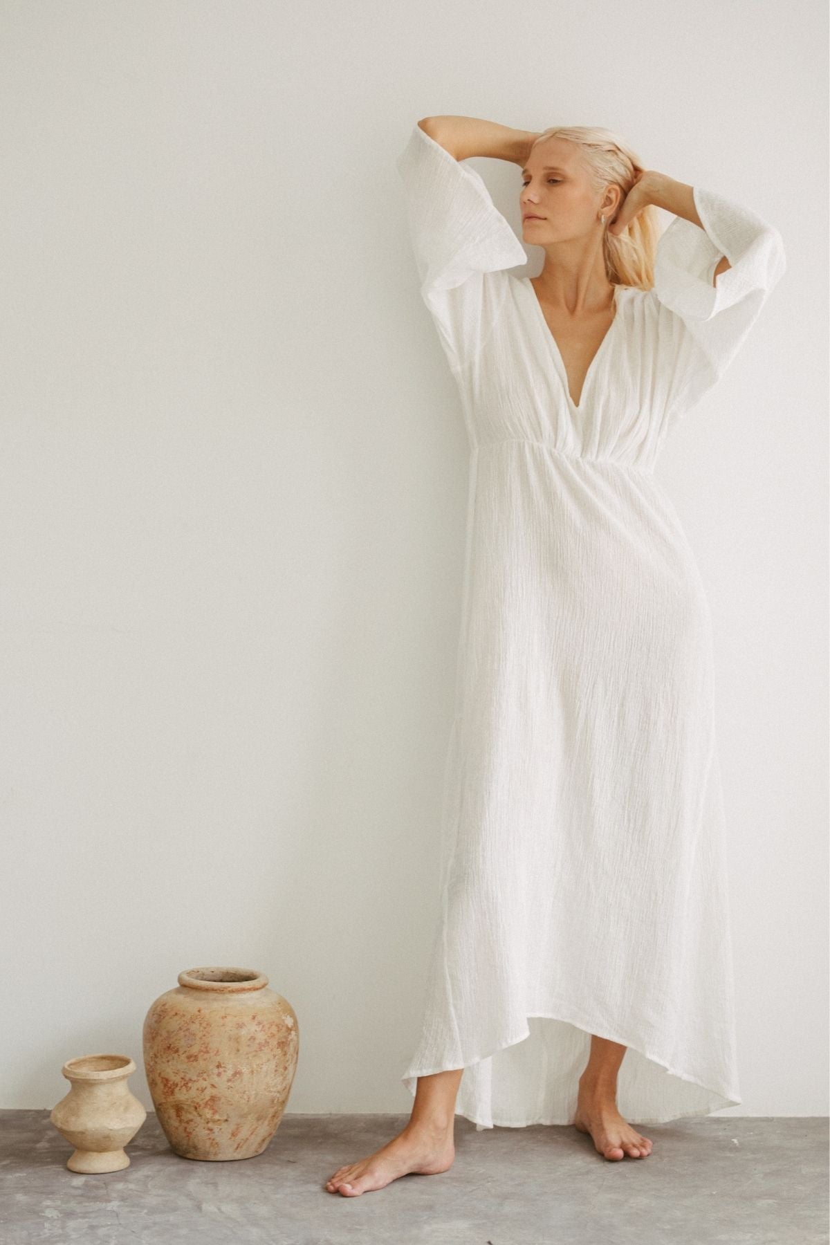 A woman with long blonde hair stands barefoot against a white wall. She is wearing the elegant Sayan Gown (Linen/Cotton) by Myrah Penaloza, featuring wide sleeves and an open back. Her arms are raised, with her hands resting on her head. Two rustic ceramic vases are placed on the floor beside her.