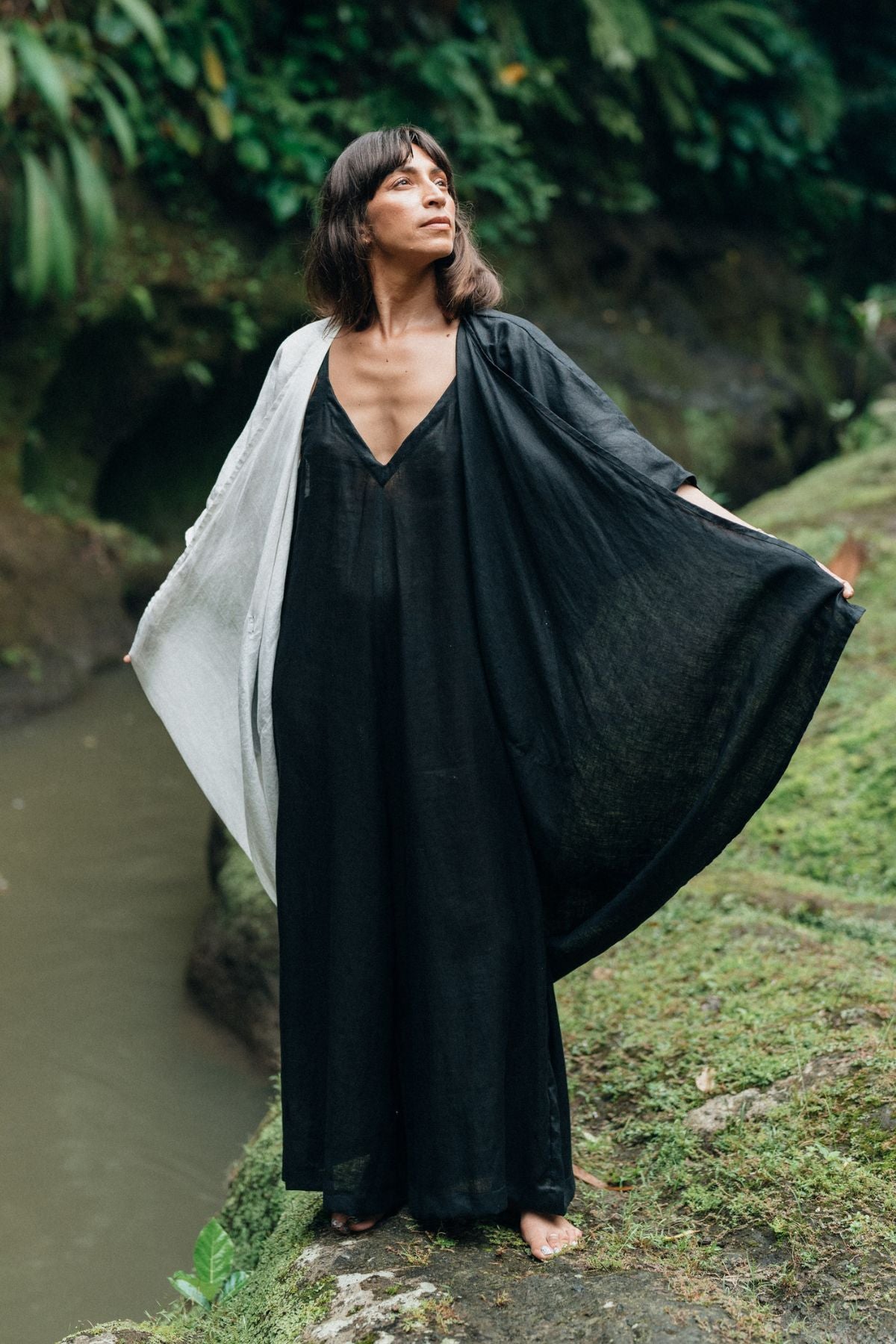 Near a river, someone is standing outdoors in a long, flowing Dao Robe from Myrah Penaloza. The robe resembles the Yin Yang design, paired with a white shawl. Their arms are outstretched, grasping the robe's sides with lush green foliage and rocks completing the background scene.