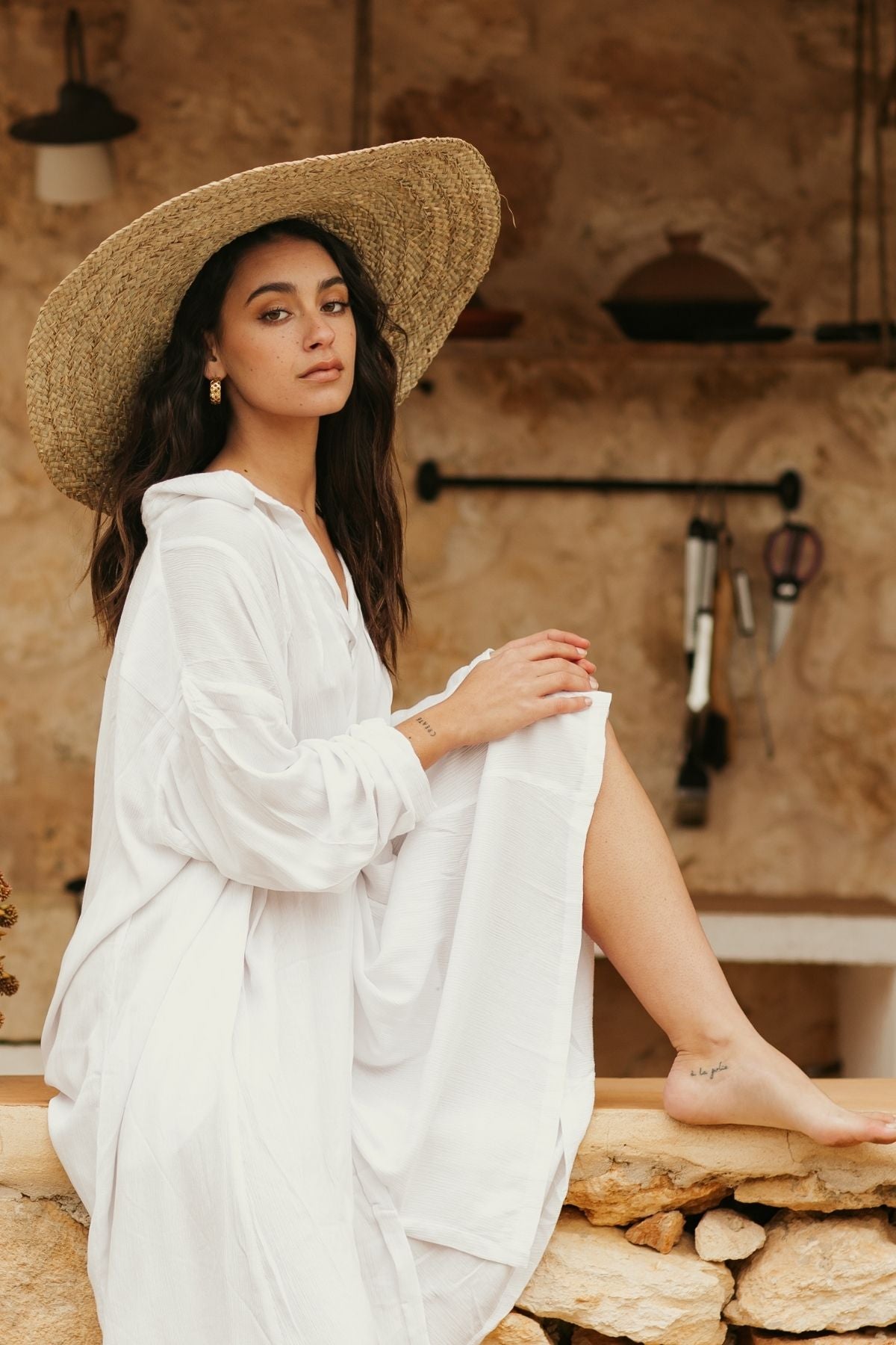 A woman in an off-white Kundalini Gown Short, made from 100% bamboo rayon by Myrah Penaloza, and wearing a large straw hat sits on a stone ledge. With a relaxed expression, she looks directly at the camera amid rustic kitchen tools that hang on the stone wall, creating a sense of elegant simplicity.