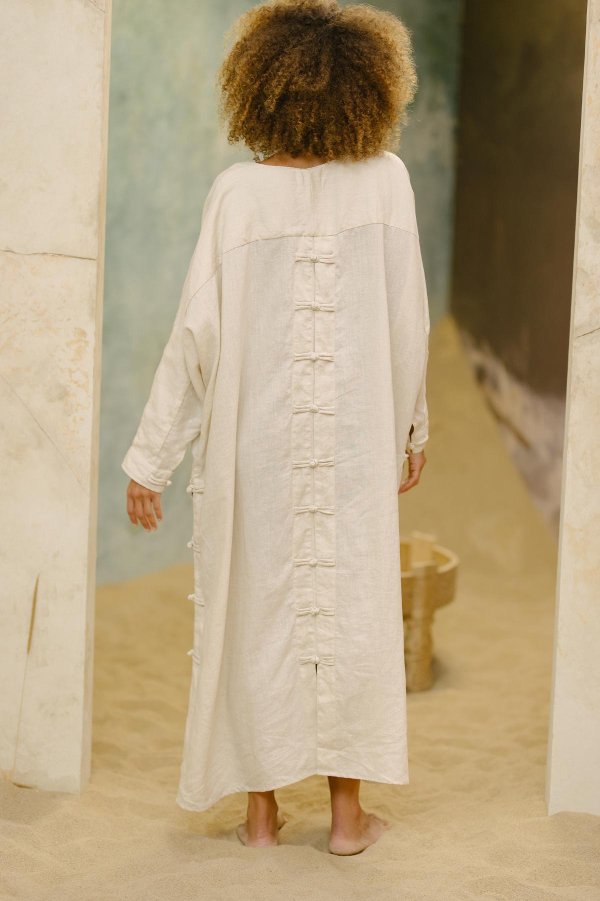 A person with curly hair, standing barefoot on sandy ground, is dressed in a long, loose beige garment. The tranquil and earthy atmosphere complements the Ancient Chajin Jacket, made to order by Myrah Penaloza, which features a row of Japanese-inspired buttons on the back.