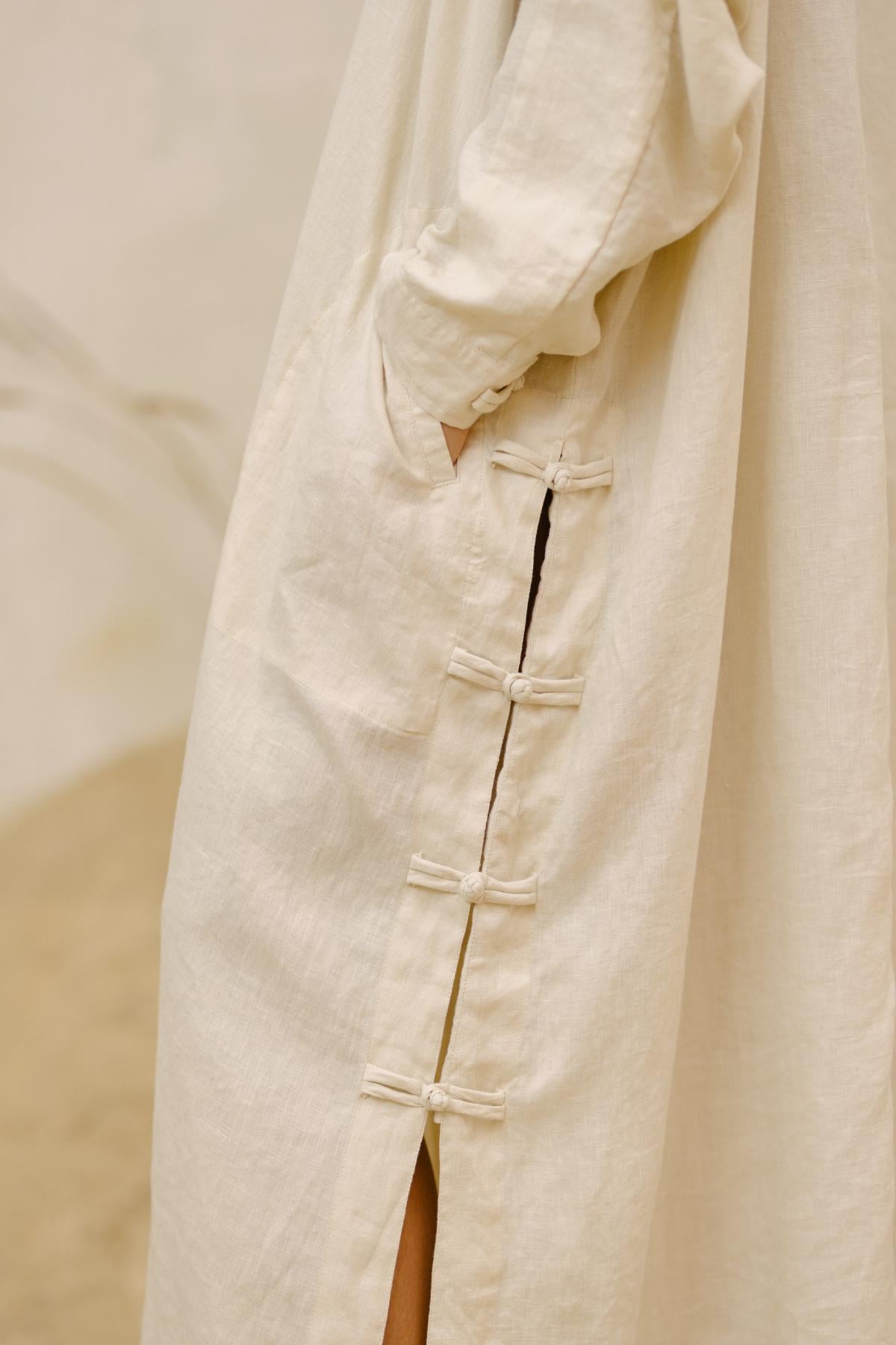 Close-up of a person wearing the Ancient Chajin Jacket (100% Linen)- Made To Order by Myrah Penaloza. The light beige, long-sleeved jacket features intricate Japanese-inspired button detailing along the side. Made from stone-washed linen, it has a pocket and a subtle texture that complements its simple, elegant design. The background is blurred and neutral-toned.