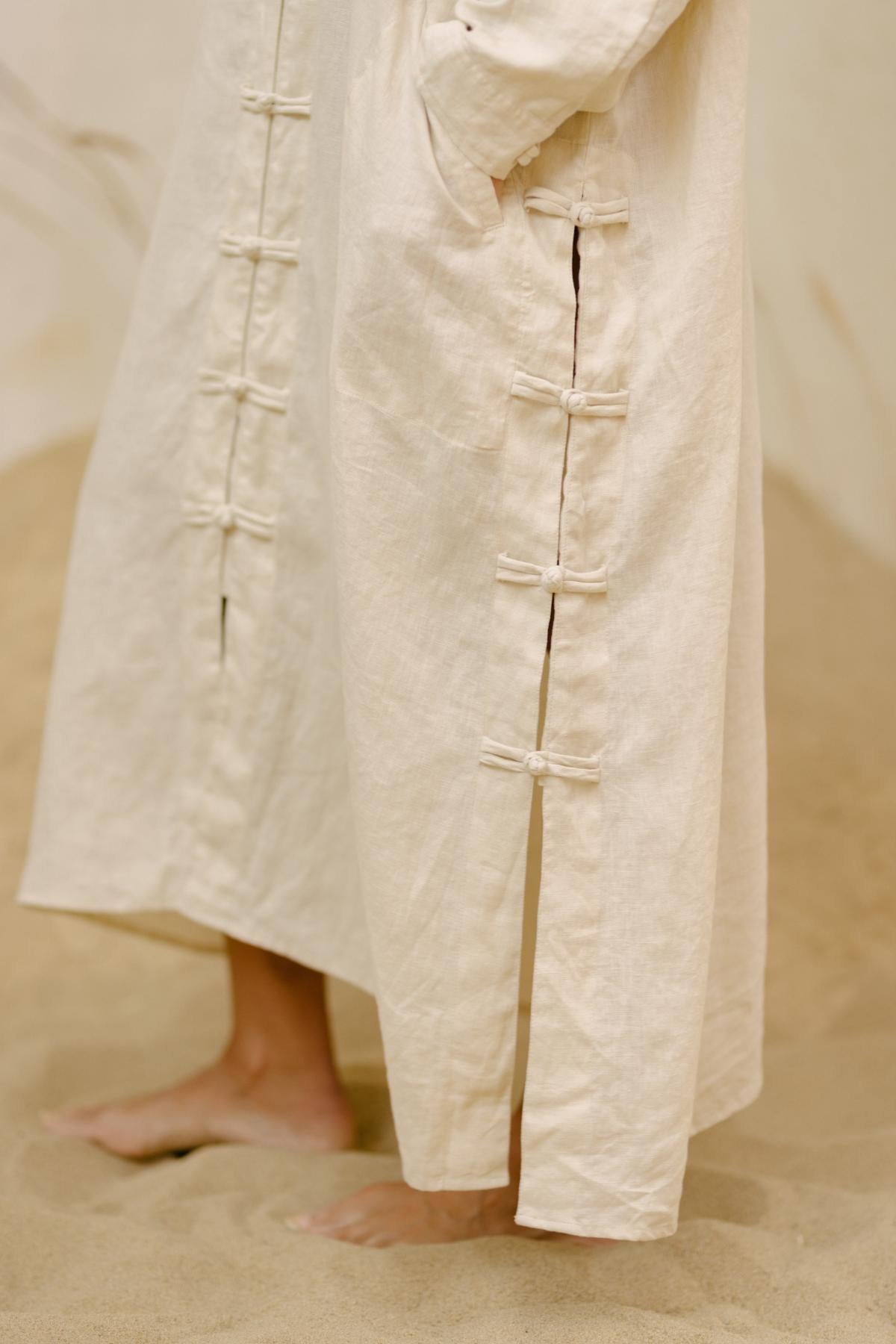 A person standing barefoot in the sand, wearing the Ancient Chajin Jacket by Myrah Penaloza. The jacket is crafted from textured, cream-colored 100% linen, featuring Japanese-inspired buttons and a slit. The long sleeves add elegance to this stone-washed look. The upper body of the person is not visible in the image.