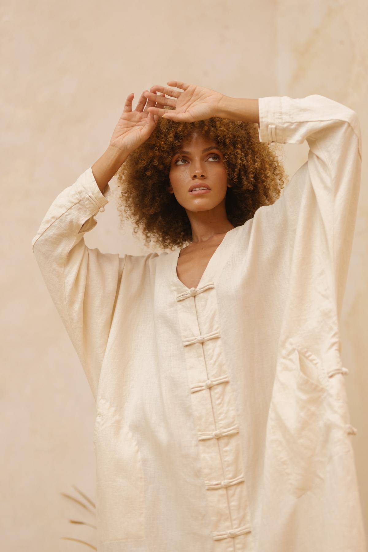A woman with curly hair strikes a pose with her arms raised, donning the Ancient Chajin Jacket from Myrah Penaloza. The cream-colored, stone-washed, 100% linen jacket, made to order, features a V-neck design accented with Japanese-inspired buttons and large pockets. Set against a soft beige background, her thoughtful expression complements the elegant simplicity of her attire.