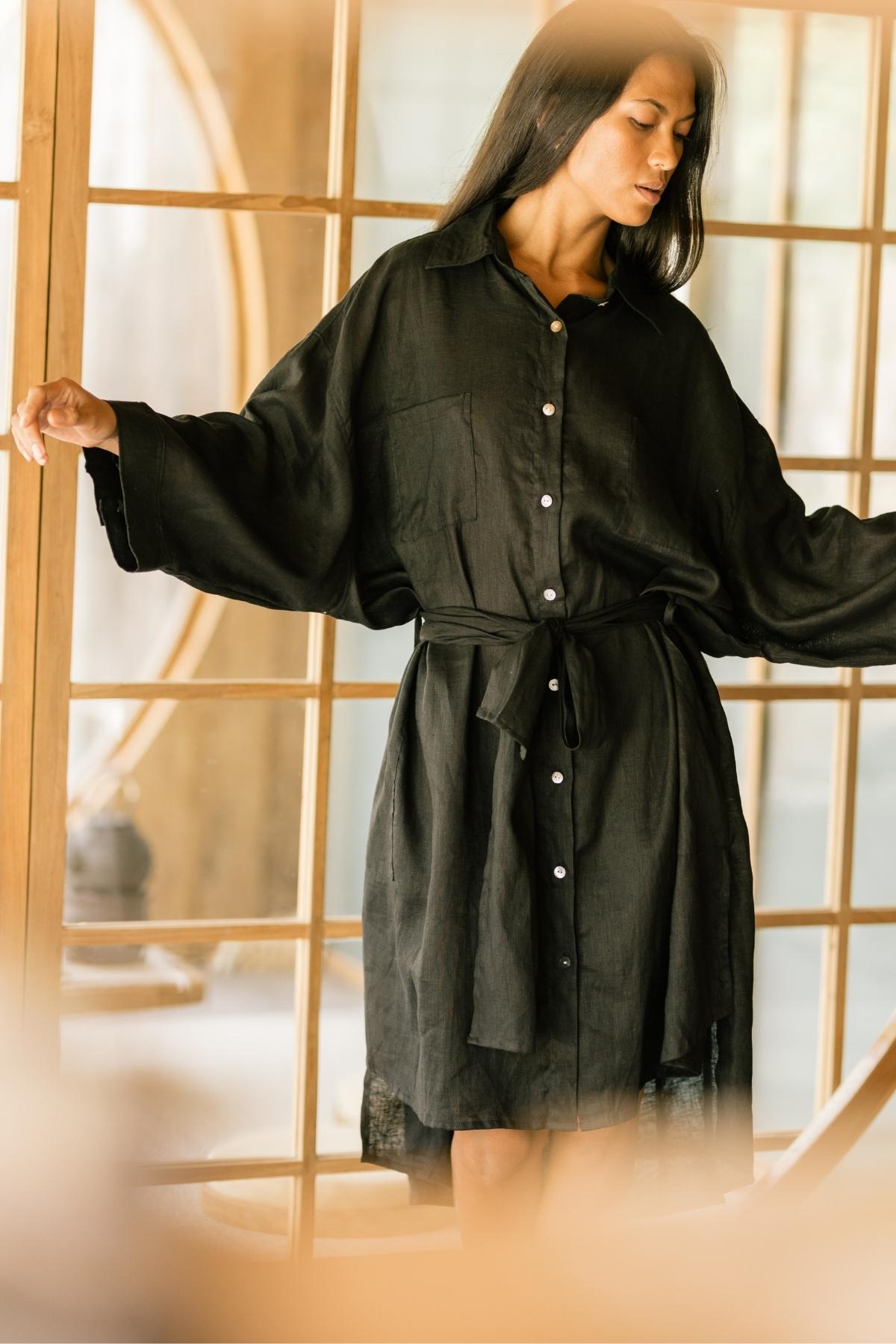 A person models the Nidra Full Length Linen Duster by Myrah Penaloza, standing gracefully in front of a wooden framed glass door with their arms slightly extended. The oversized fit drapes elegantly under the warm indoor lighting, creating a calm and serene atmosphere that emphasizes the elegance of sustainable fashion.