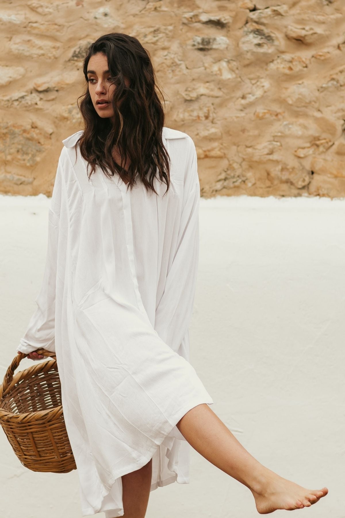 In a loose, off-white Kundalini Gown Short by Myrah Penaloza, fashioned from 100% bamboo rayon, a woman stands barefoot on a white surface with a wicker basket in hand. Her long, wavy hair cascades down as she gazes thoughtfully, complemented by the rustic charm of the textured stone wall behind her.