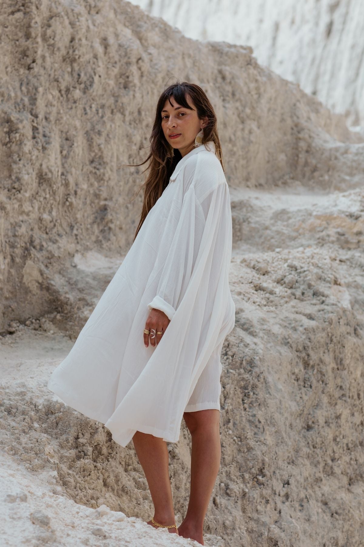 A woman in an off-white Kundalini Gown Short crafted from 100% bamboo rayon by Myrah Penaloza stands on rugged terrain. She gazes over her shoulder with a serene expression, her dark hair gently swaying in the wind. The textured, light-colored rock formations in the background highlight the elegance of her dress.
