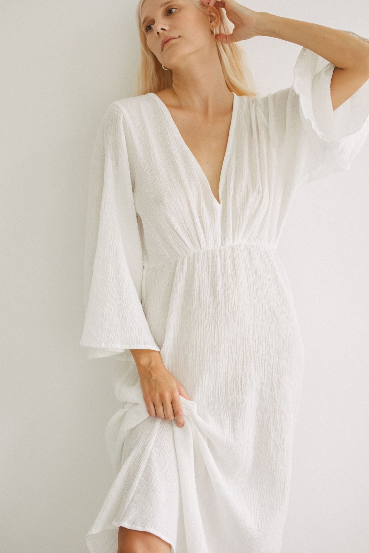 A woman with blonde hair is wearing the Sayan Gown, a long-sleeve, V-neck dress made of textured linen and cotton from Myrah Penaloza. She is posing against a plain white background, gently touching her ear with one hand and holding the hem of her elegant open-back gown with the other.