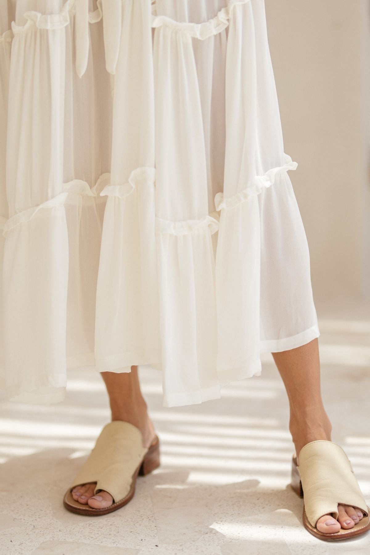 A person stands on a light-colored floor wearing beige mules and the flowing La Dama skirt by Myrah Penaloza, crafted from 100% silk. The sleeveless design with a ruffle neck exudes a renaissance gown feel, as the soft fabric drapes gracefully and light creates subtle patterns on its tiers.