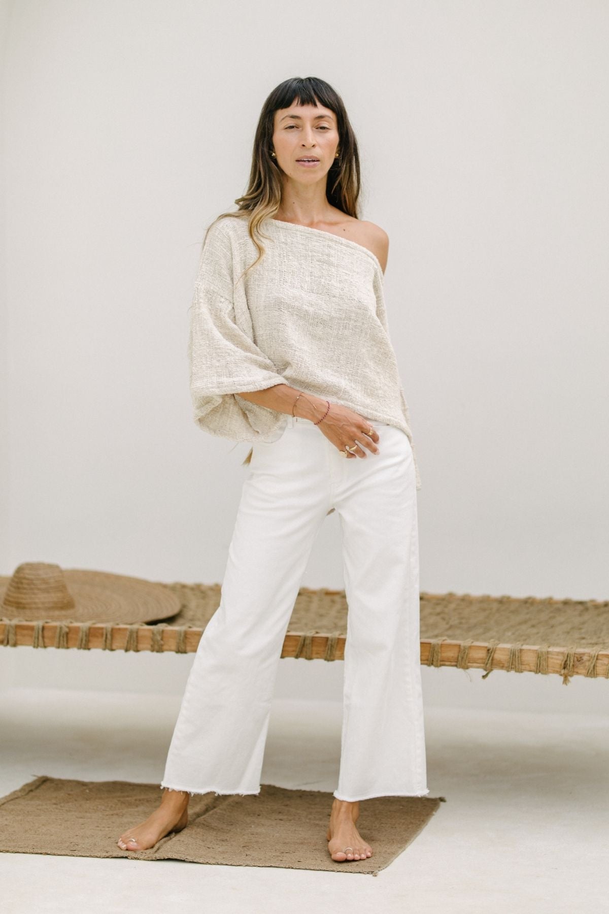 A woman stands barefoot indoors against a neutral background, dressed in a loose, off-the-shoulder beige top and Myrah Penaloza's comfortable Miley (High Waisted) Jean - Pre Order Only. She is standing on a natural woven rug, with a woven mat and a straw hat in the background. Her relaxed pose exudes a casual vibe.