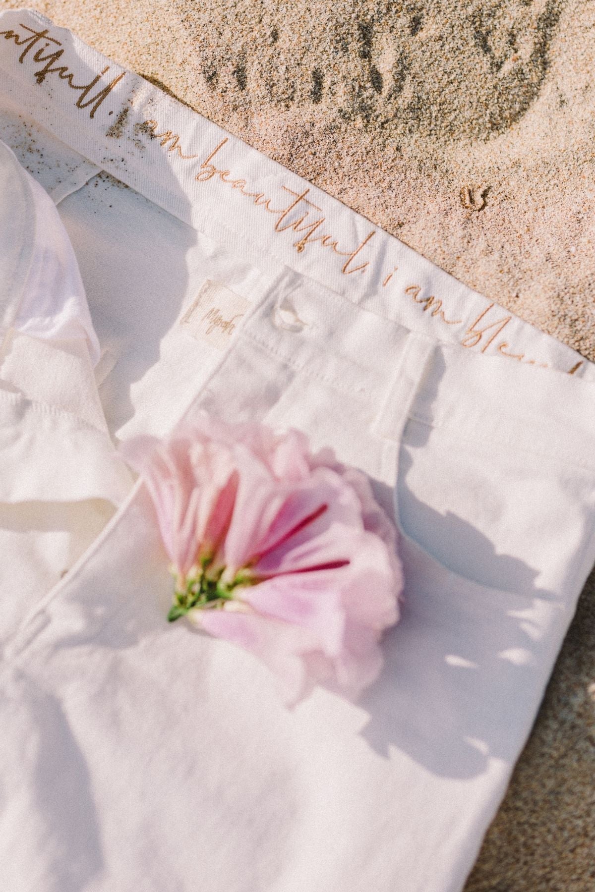 A pair of Miley (High Waisted) Jeans from Myrah Penaloza lies on a sandy surface, with a pink flower placed over the waistband. There is cursive text on the waistband that appears to read "I am beautiful" and "I am blessed." Shadows and sunlight highlight the scene, indicating a bright, sunny day. Note: This item is available for pre-order only.