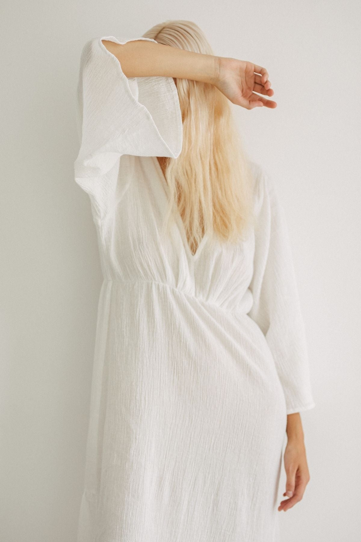 A person with long blonde hair, dressed in an elegant Sayan Gown by Myrah Penaloza, poses against a plain background. Their arm is raised, covering part of their face, while the other hand rests on their hip. The gown features wide sleeves and a slightly gathered waist and is meticulously crafted from linen and cotton.