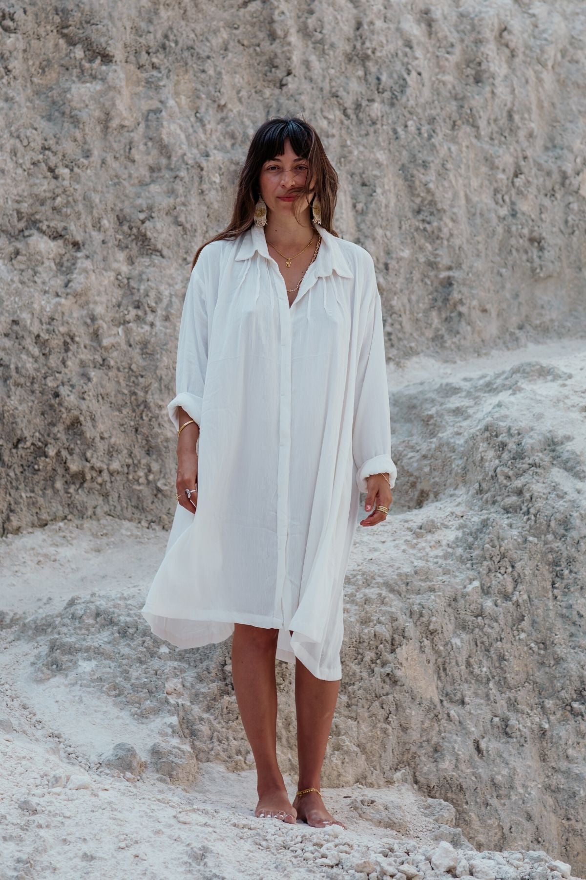 Dressed in a flowing Kundalini Gown Short by Myrah Penaloza, made entirely from soft bamboo rayon in an off-white hue, a person stands barefoot on rocky, sandy terrain. The backdrop features a textured, light-colored rock formation. With long hair and facing the camera, they capture the serene essence of this natural setting.
