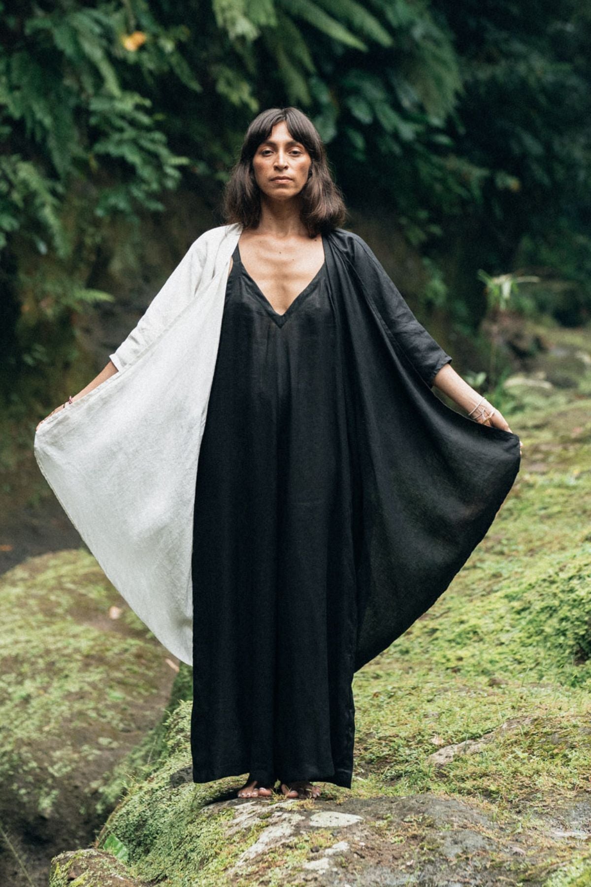 A woman stands outdoors on moss-covered rocks, wearing a flowing Dao Robe by Myrah Penaloza in a yin-yang design, crafted from 100% linen. She holds the sides of the robe outspread, with lush greenery in the background.