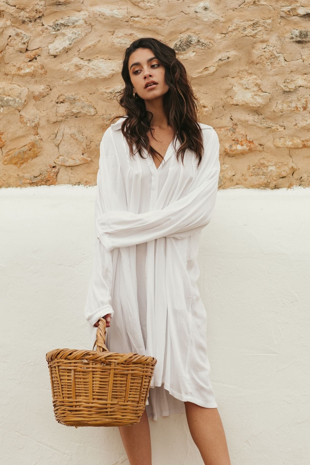 A woman in an off-white Kundalini Gown Short by Myrah Penaloza leans against a white and stone wall, holding a wicker basket. Her long, wavy hair cascades gracefully as she stands with a relaxed posture, embodying the effortless elegance of handmade garments crafted from 100% bamboo rayon.