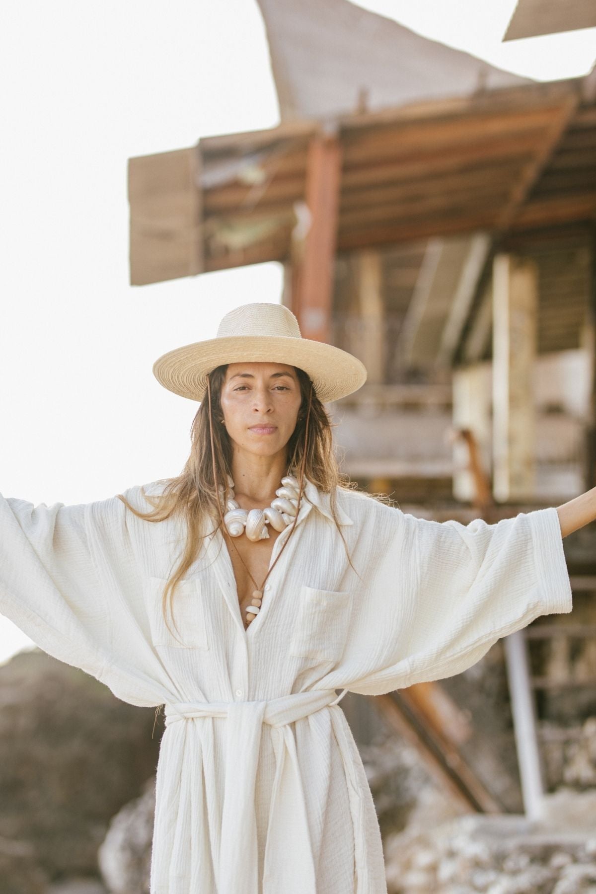 Wearing a white robe and a wide-brimmed hat, a woman adorned with Myrah Penaloza's Aphrodite's Treasure Necklace poses with her arms outstretched. She stands before a rustic wooden structure set against a rocky landscape, illuminated by the bright, sunlit backdrop.