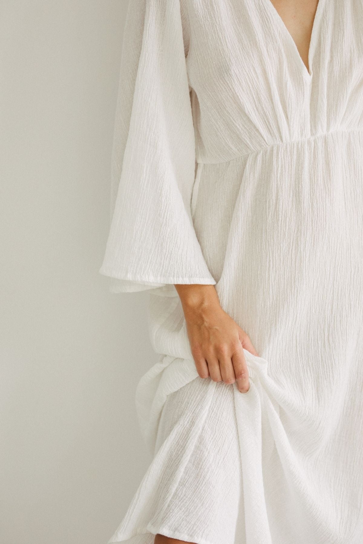 A person wearing the Sayan Gown (Linen/Cotton) by Myrah Penaloza gently grasps the textured, white, long-sleeve fabric of the dress with one hand. The image focuses on the torso and intricate details of the gown, showcasing artisanal craftsmanship typical of handmade tailors, with the person's face not visible.