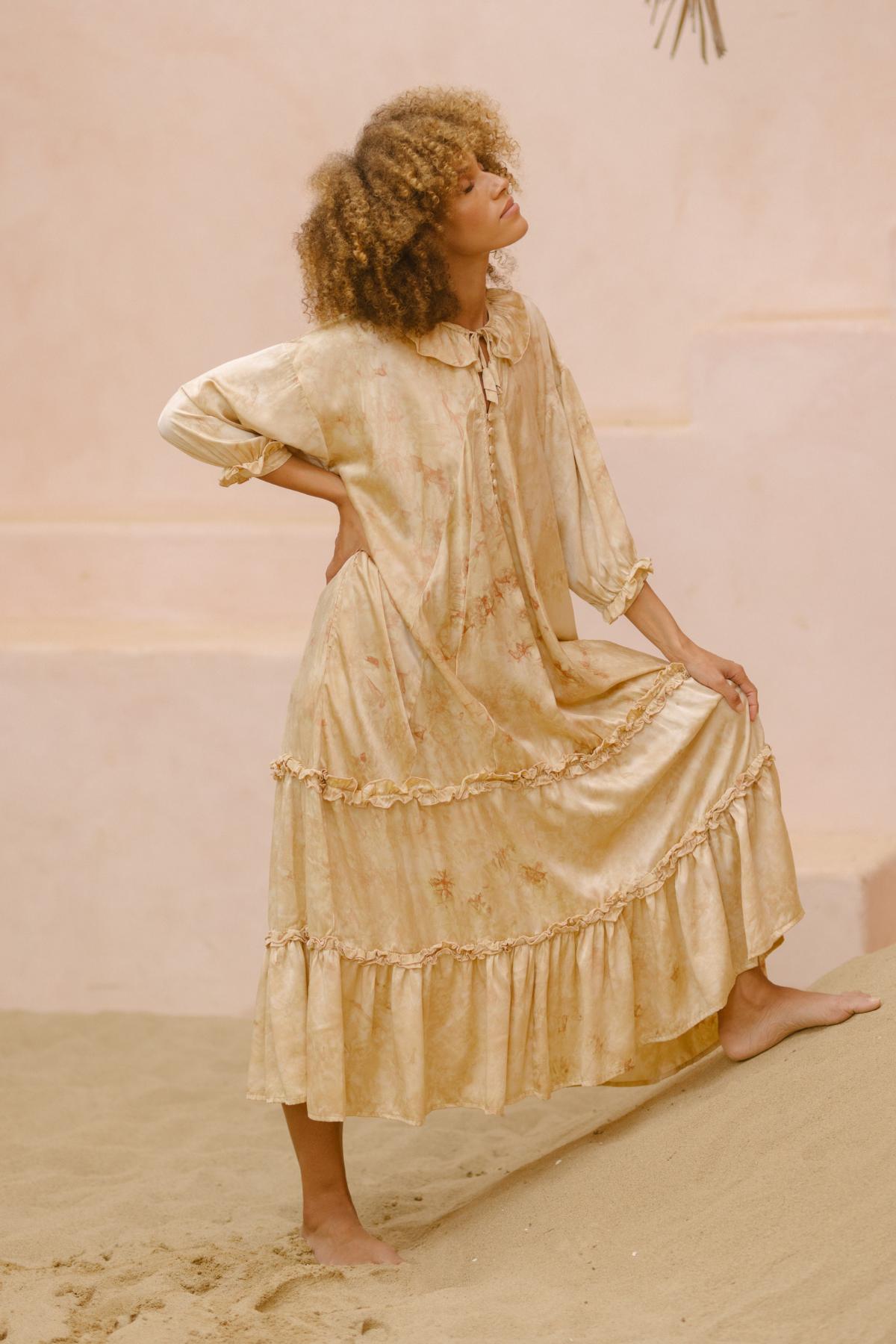 A person with curly hair is posing outdoors in the Allegra Joy, Rose Petal Dyed, Silk Gown - Made To Order by Myrah Penaloza. The flowing beige dress features ruffles and a floral pattern. They stand on one bent leg, with one hand on their hip and the other holding up part of the vintage couture gown. The background features a sandy surface and muted tones.