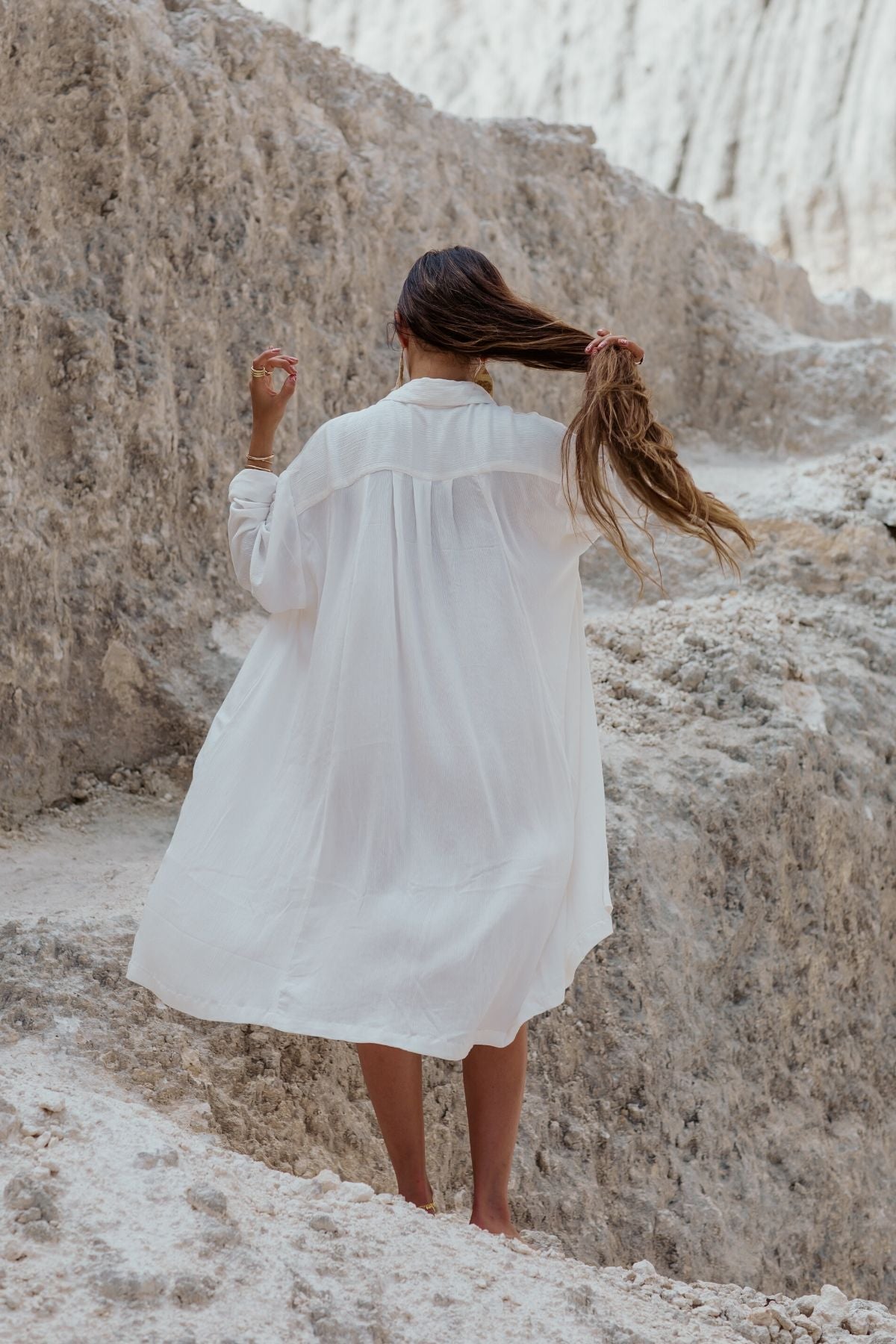A woman in a flowing Myrah Penaloza Kundalini Gown Short, made from 100% bamboo rayon in off-white, stands on a rocky terrain with her long hair catching the breeze. The rugged and sandy landscape enhances the serene natural ambiance, perfectly reflecting the elegance of handmade garments crafted from bamboo rayon.