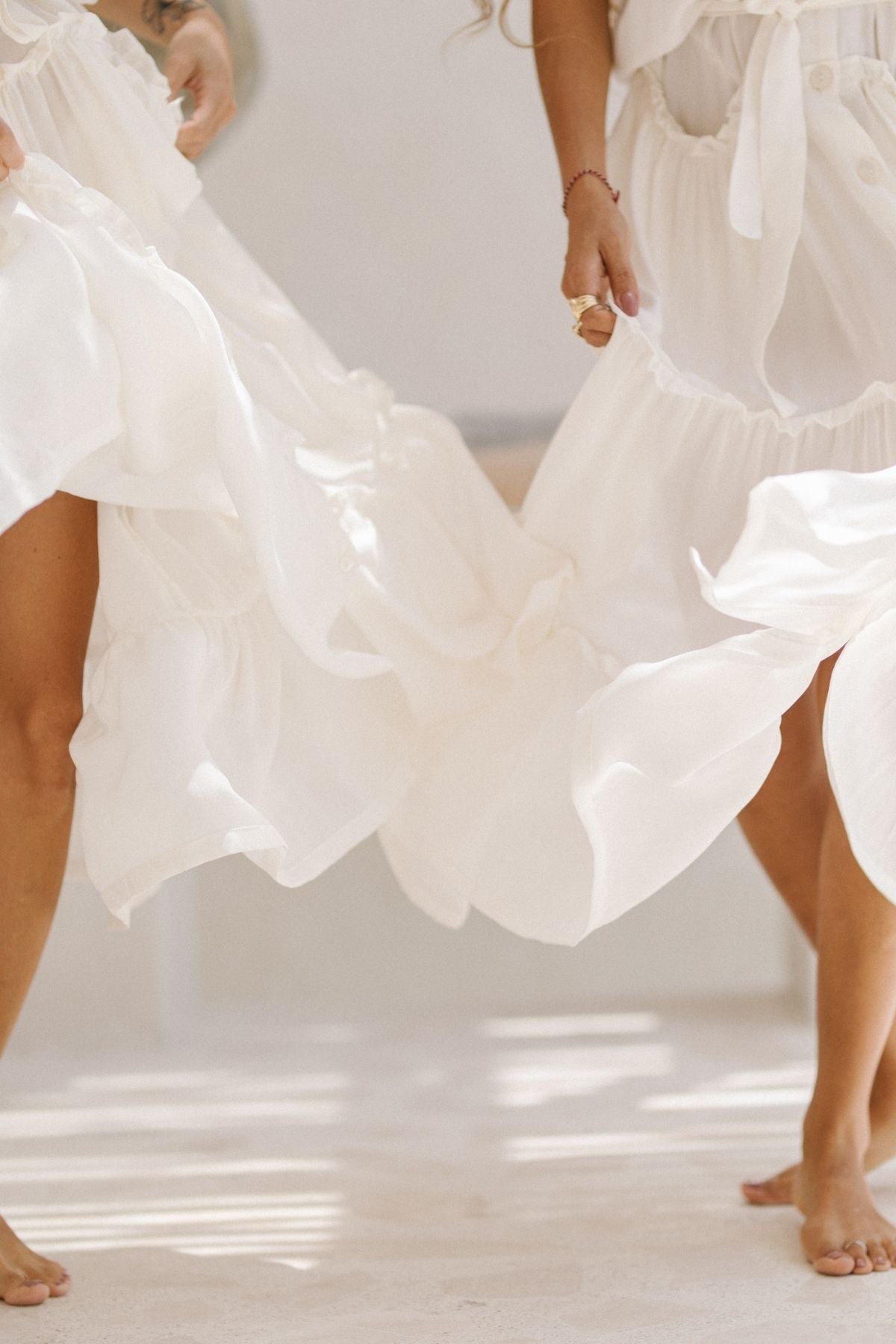 Two individuals in flowing, white La Dama dresses from Myrah Penaloza, reminiscent of a renaissance gown, dance barefoot on a light-colored floor. The 100% silk fabric of the sleeveless dresses with ruffle necks is in motion, creating a sense of elegance. Sunlight casts soft, warm shadows on the scene, highlighting the grace of their movements.