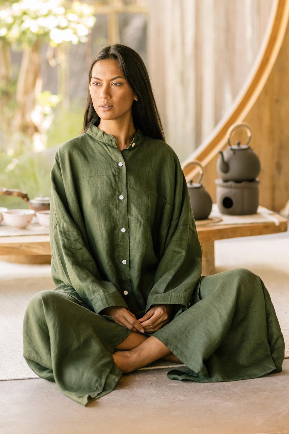 A person with long dark hair sits cross-legged on the floor, donning a loose-fitting, long-sleeved Camisa Del Novio Linen top and Shanti Skirt Set from Myrah Penaloza. Behind them, a wooden table with a teapot set enhances the serene ambiance, complemented by lush greenery in the background. The 100% linen fabric adds to the tranquil atmosphere of the scene.