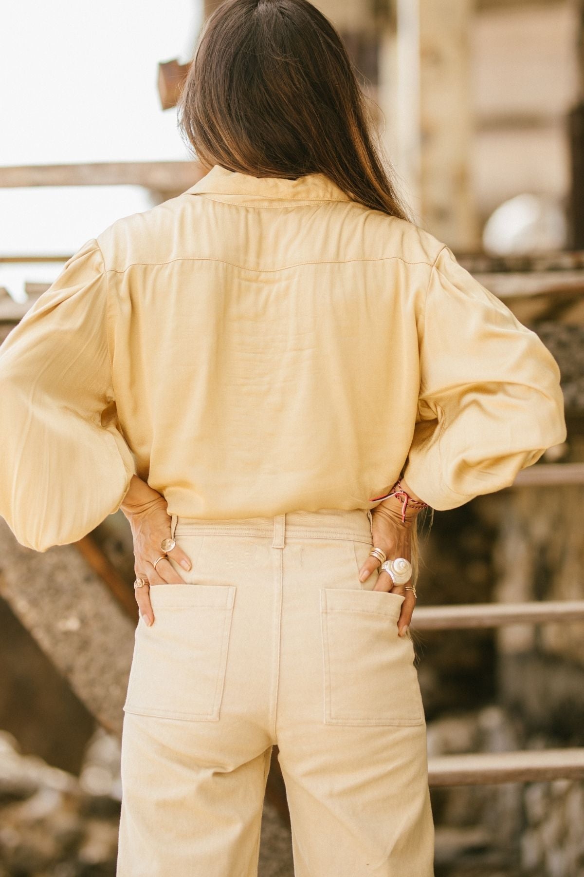 A person with long hair stands with their back to the camera, dressed in a light beige long-sleeve blouse and Myrah Penaloza's Vida high-waisted stonewashed jeans, botanically hand-dyed. Their hands rest on their hips, showcasing rings and bracelets, against a backdrop of stairs and a blurred outdoor setting.