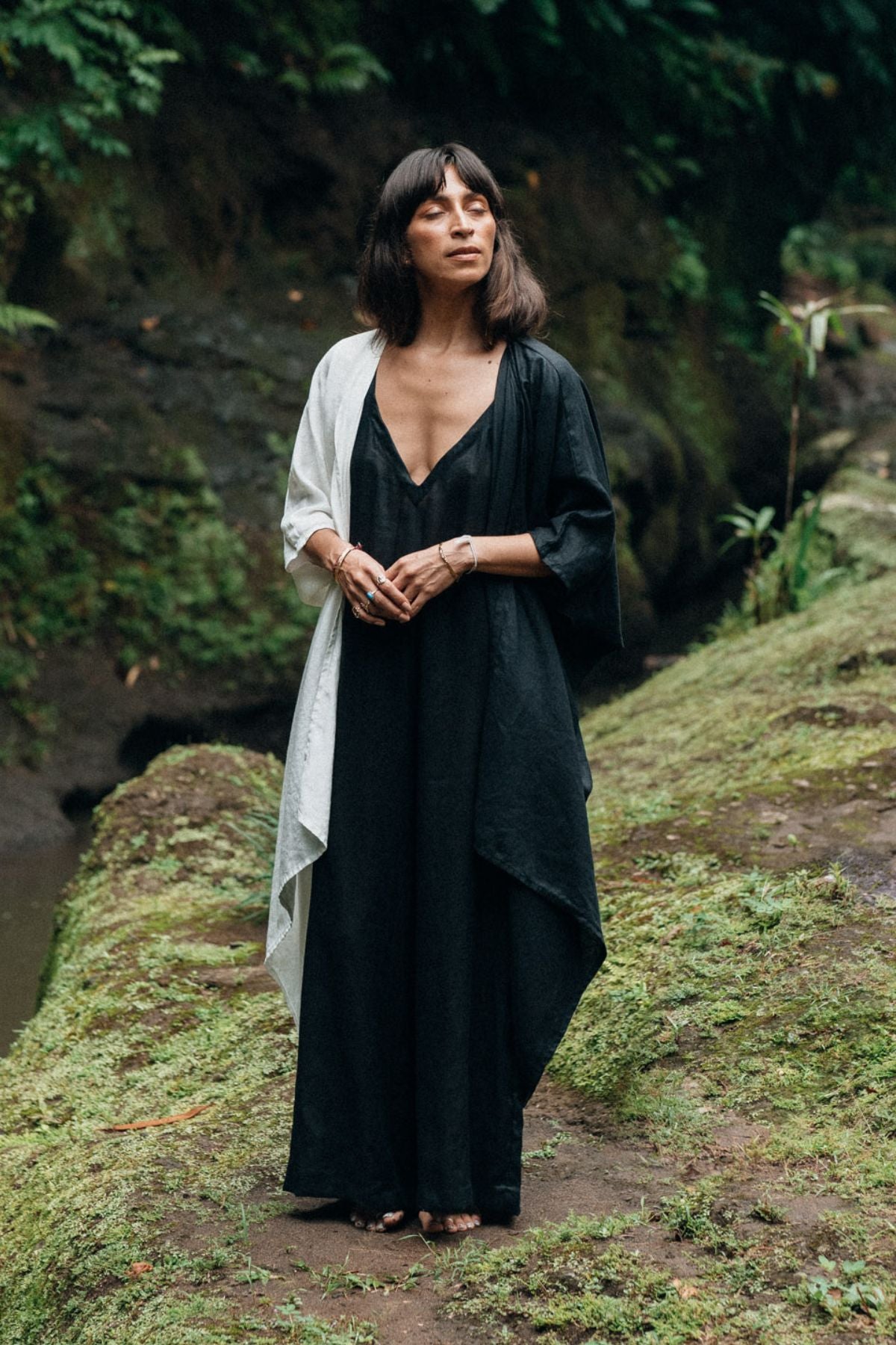 Standing on a mossy path surrounded by lush greenery, a person dons a Myrah Penaloza Dao Robe (Yin Yang, Set Optional, Made To Order), exuding the elegance of a long black dress. A white shawl drapes gracefully over one shoulder as they stand with eyes softly closed, embodying serenity in the tranquil setting.