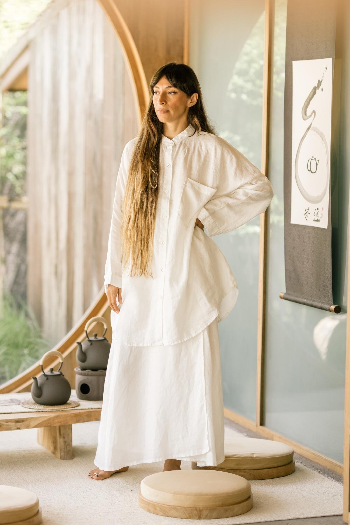 A woman stands in a minimalist room with circular wooden features, dressed in Myrah Penaloza's Camisa Del Novio Linen Top and Shanti Skirt Set. She has long straight hair with bangs. In the background, there is a hanging scroll with artwork, and a low wooden table features a black teapot and cups. Cushions are arranged on the floor.