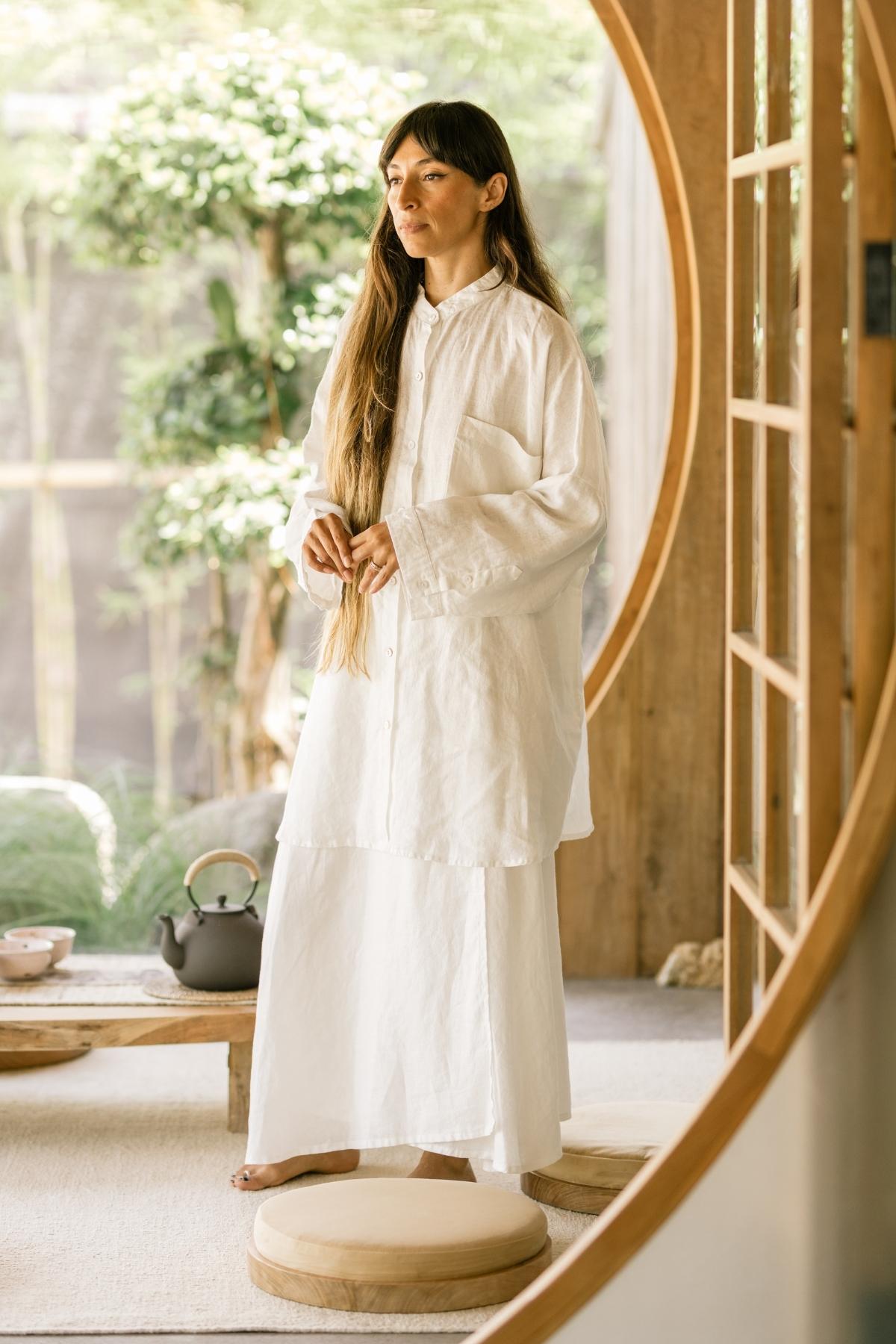 A woman stands in a tranquil room with a circular wooden doorway and lush greenery visible outside. She wears the Myrah Penaloza Camisa Del Novio Linen top paired with the Shanti Skirt, and her long hair cascades down her back. A tea kettle and cups sit on a low wooden table next to round cushions on the floor.