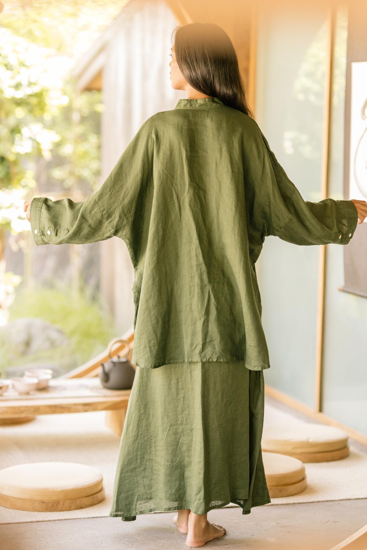 A woman with long dark hair, seen from the back, stands barefoot indoors in a softly lit room. She is wearing a loose-fitting, olive-green Camisa Del Novio Linen top paired with the Shanti Skirt from Myrah Penaloza. The room features wooden furniture and a large window showcasing greenery outside.
