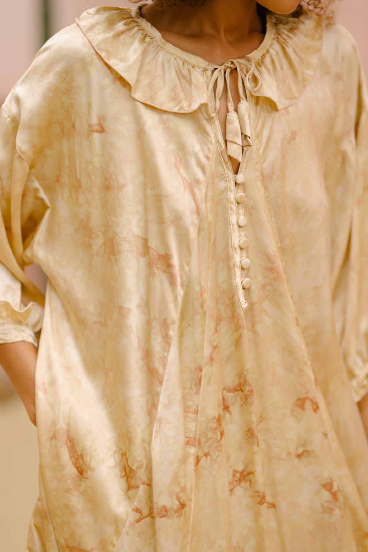 A person wearing the Allegra Joy, Rose Petal Dyed, Silk Gown by Myrah Penaloza, which features a light beige hue with a ruffled neckline and string tie closure. The gown is adorned with small buttons down the front and showcases subtle, soft patterns in shades of light brown. The person's hands rest in pockets on either side.
