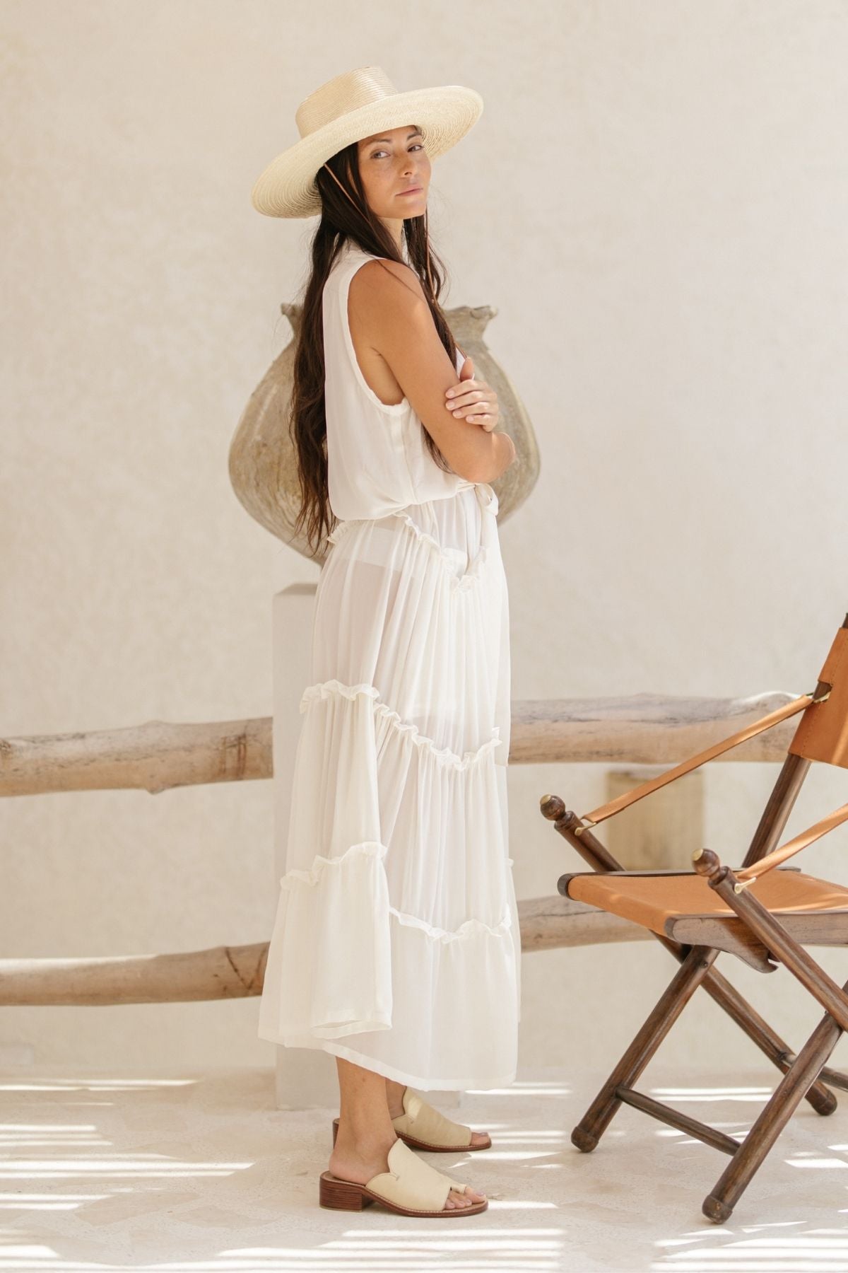 In a light, airy room, a woman with long hair stands sideways, wearing the La Dama – a sleeveless, ruffle neck dress crafted from 100% silk by Myrah Penaloza. Accompanied by sandals and a wide-brimmed hat, she stands beside a wooden chair and rustic clay pot against the neutral backdrop, effortlessly exuding elegance reminiscent of a renaissance gown.