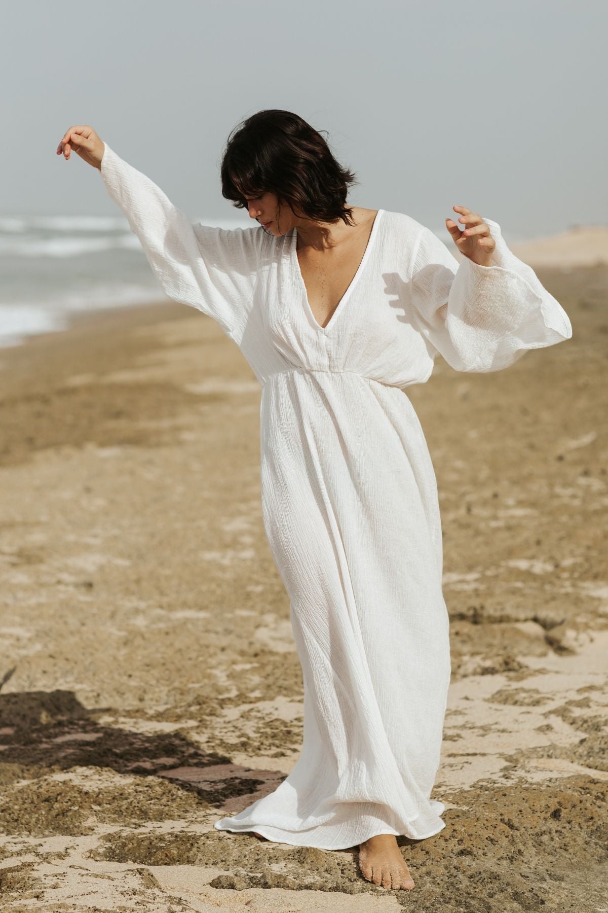 A person in a Sayan Gown by Myrah Penaloza dances barefoot on a sandy beach. They have short, dark hair and are illuminated by natural light. The ocean waves and a hazy sky can be seen in the background.