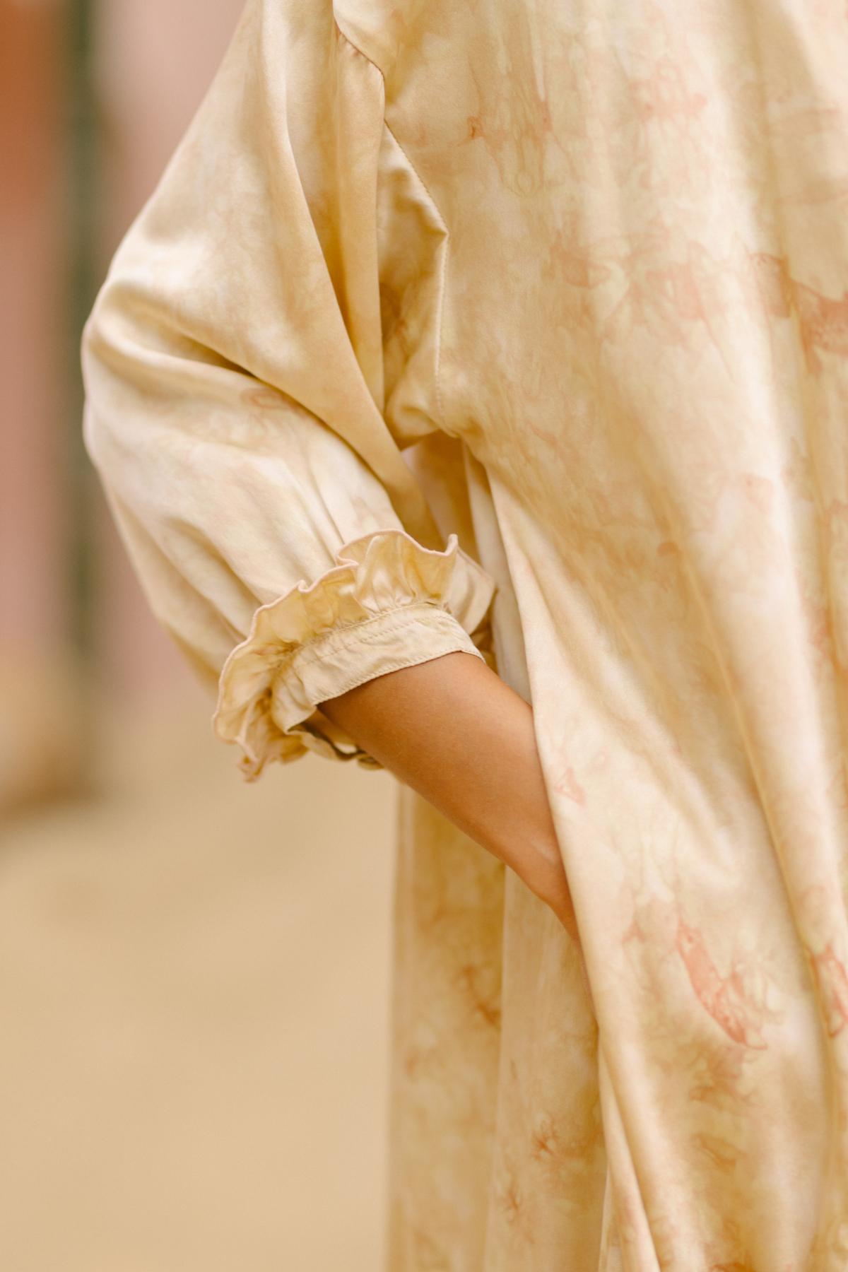 A close-up of someone wearing the Allegra Joy gown by Myrah Penaloza, crafted from rose petal dyed silk. The loose, long-sleeved dress in soft creamy yellow features a subtle marble-like pattern. The person's hand rests in the side pocket, and delicate ruffled cuffs adorn the sleeves, evoking a vintage couture feel.