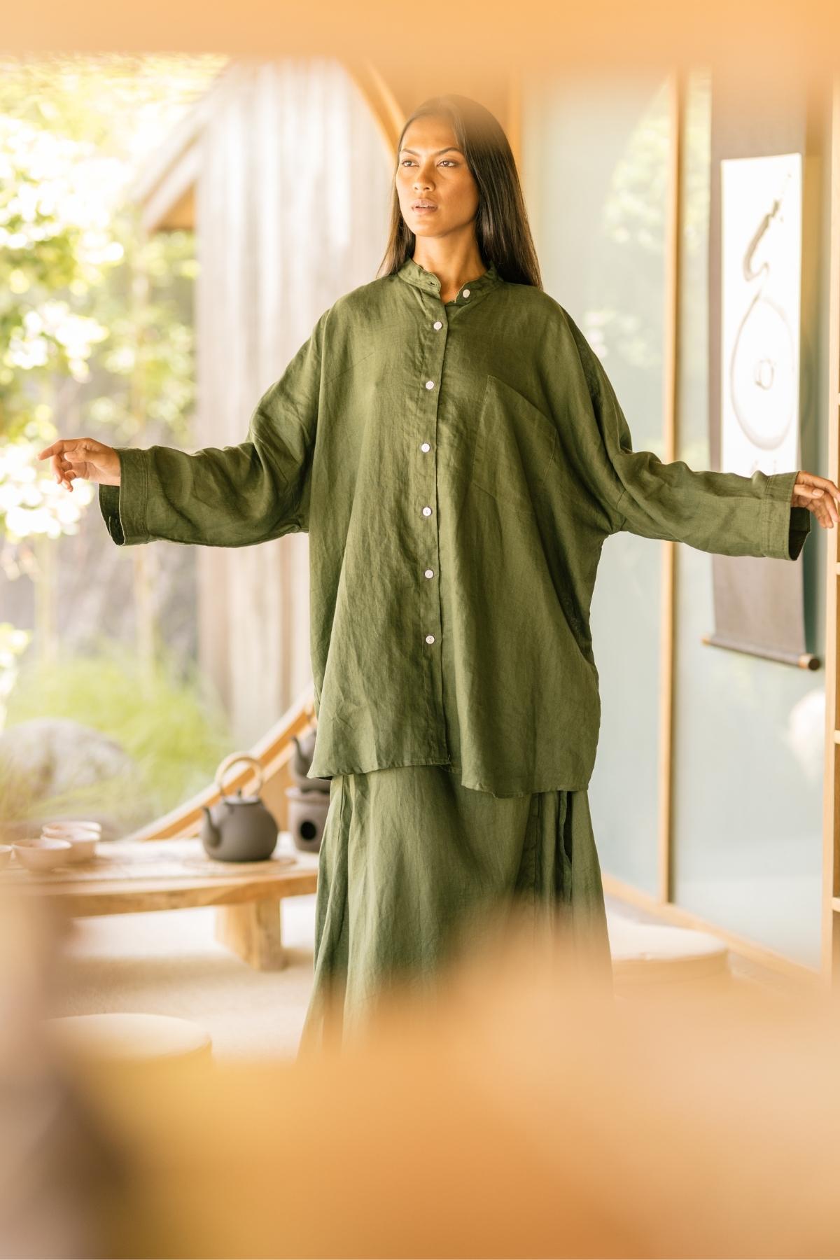 A woman wearing the Camisa Del Novio Linen top + Shanti Skirt Set by Myrah Penaloza stands with her arms slightly extended. She is in a serene, well-lit room with a tea set in the background and a scroll hanging on the wall. The setting appears calm and inviting.