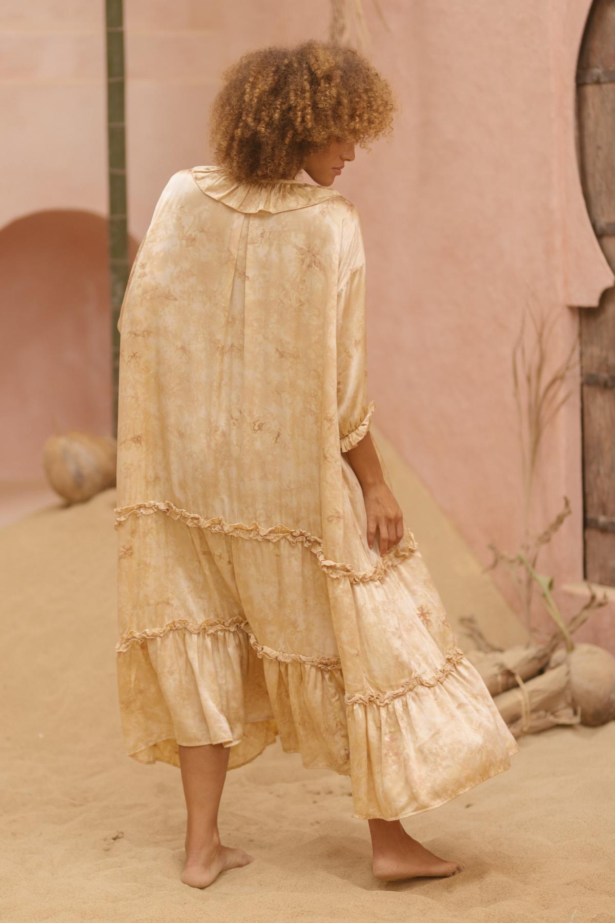 A person with curly hair stands barefoot on a sandy surface, wearing the flowing, Allegra Joy, Rose Petal Dyed Silk Gown by Myrah Penaloza. The made-to-order vintage couture gown features ruffled layers and a subtle pattern. The person is turned slightly away, showcasing the back of the garment. Nearby are rustic walls and a piece of driftwood.