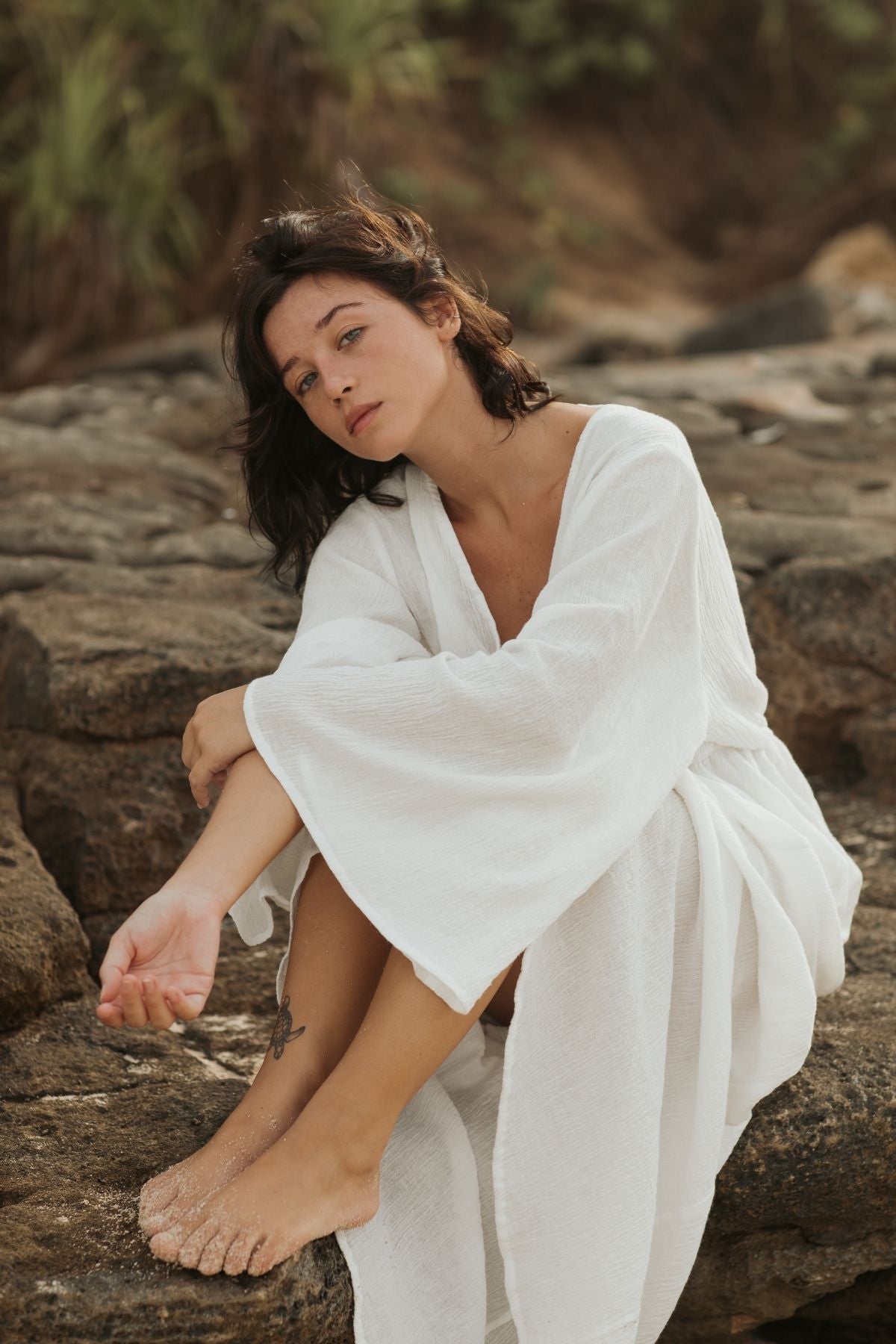 A woman with dark hair sits on rocky terrain, draped in the elegant Sayan Gown made of linen and cotton from Myrah Penaloza. Her expression is relaxed, and her feet, one of which bears a small tattoo, are covered in sand. She is surrounded by natural outdoor surroundings with green foliage in the background.