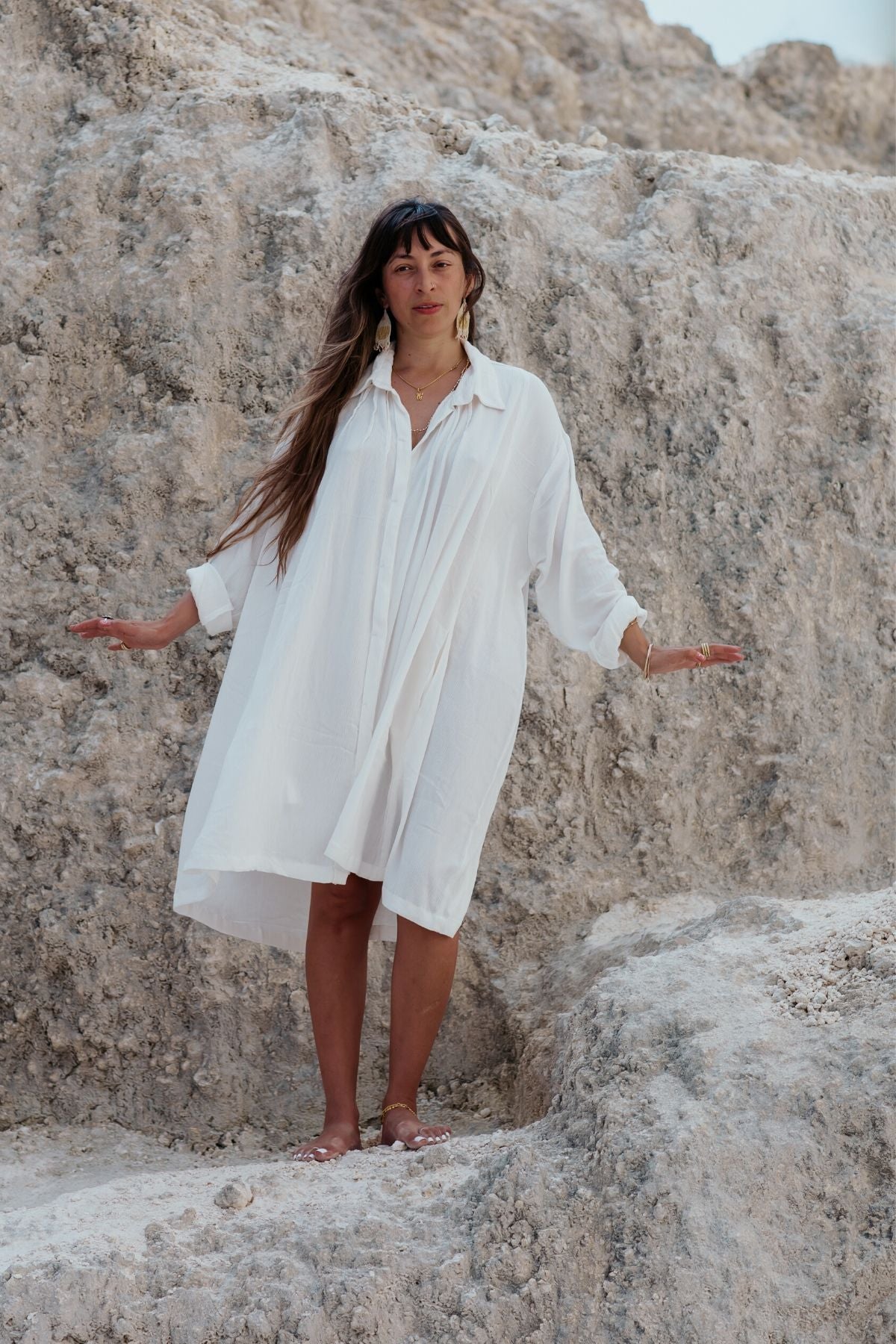 Barefoot and standing confidently on rocky terrain, a person with long hair wears the off-white, flowing Kundalini Gown Short by Myrah Penaloza, crafted from 100% bamboo rayon. They extend their arms slightly to maintain balance on the uneven ground, with a backdrop of textured stone completing the scene.