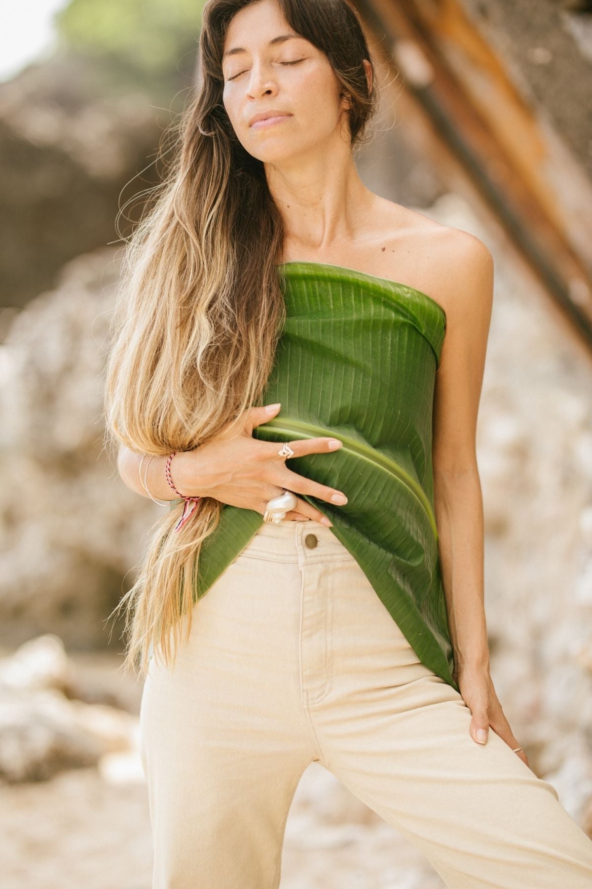 Outdoors, a woman with long, wavy hair and closed eyes stands relaxed and content against a backdrop of natural scenery. She wears a strapless top fashioned from a large green leaf and light-colored high-waisted jeans named Vida by Myrah Penaloza, which are stonewashed and botanically hand dyed.
