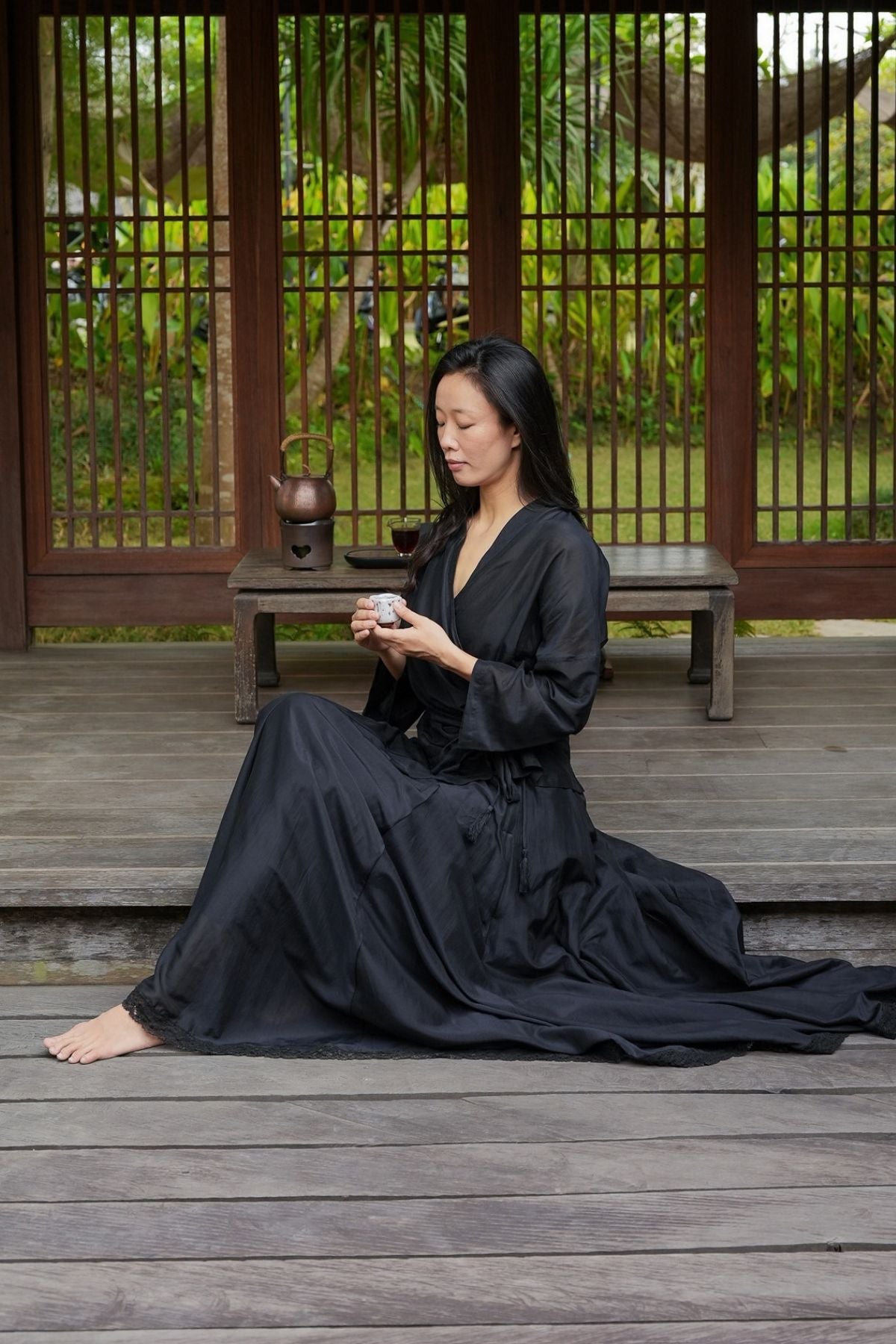 The Perla Set in Dark Moon Black silk and cotton, worn seated with a cup of tea in a wooden tea pavilion in Bali